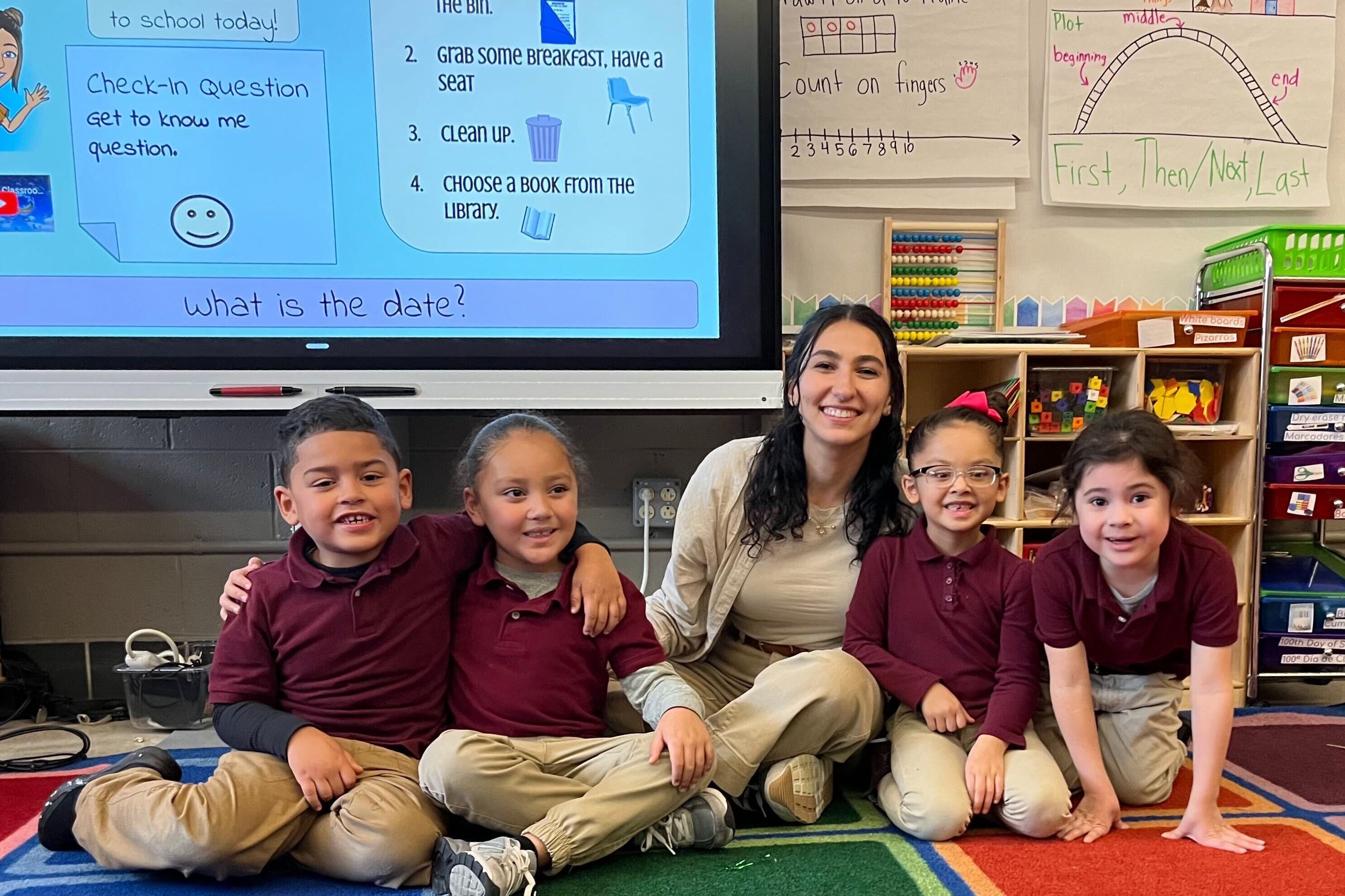 An adult woman wearing all tan sits between four young students wearing school uniforms with a projector and white board in the background.