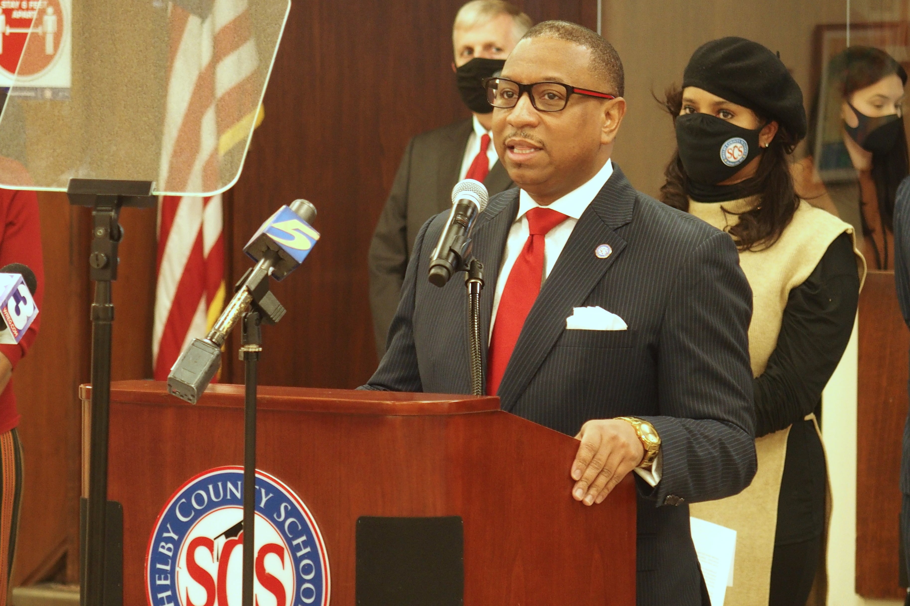 Superintendent Joris Ray stands at a podium with microphones during a press conference as school board chairwoman Miska Clay Bibbs, wearing a mask, looks on from behind.