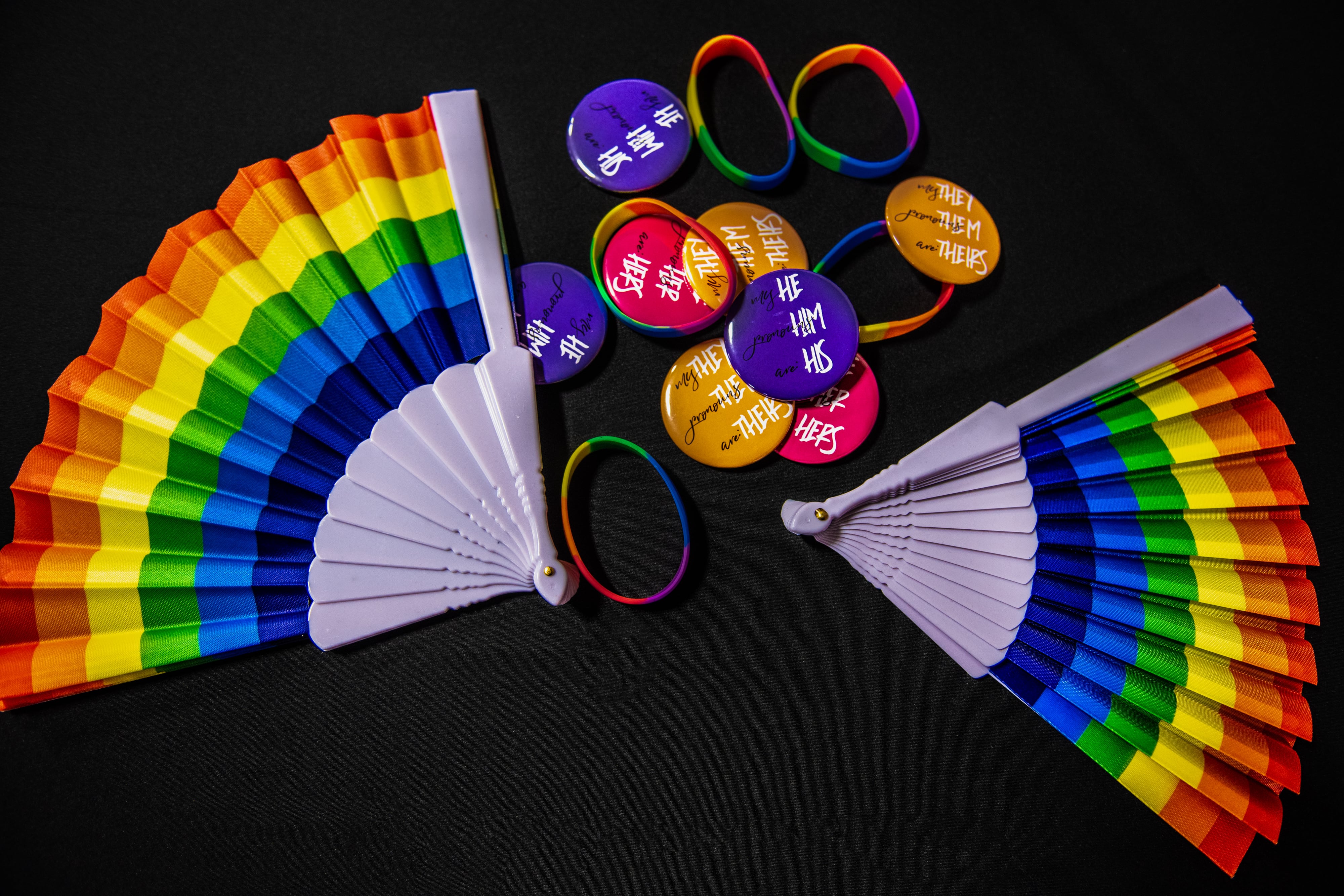 Colorful Pride fans and pins with pronouns sit on a black tablecloth.