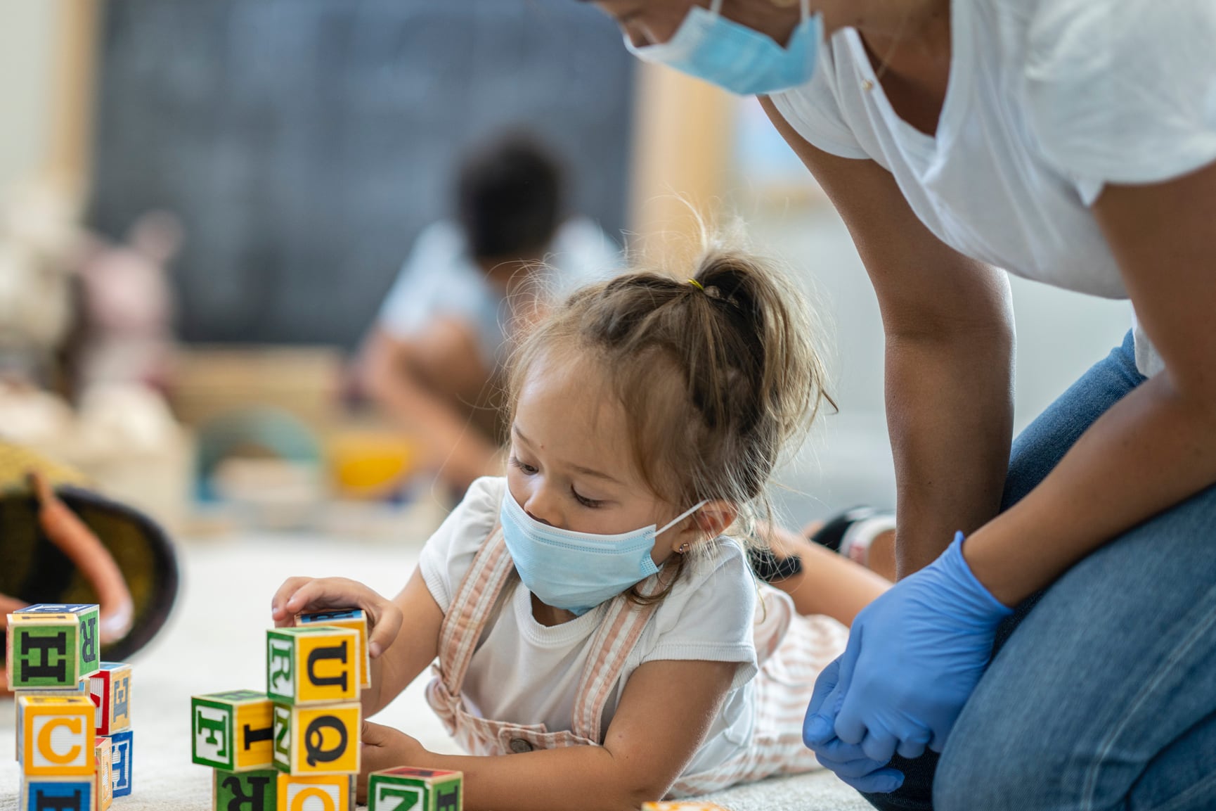 Young girl in mask plays with blocks while masked and gloved woman watches