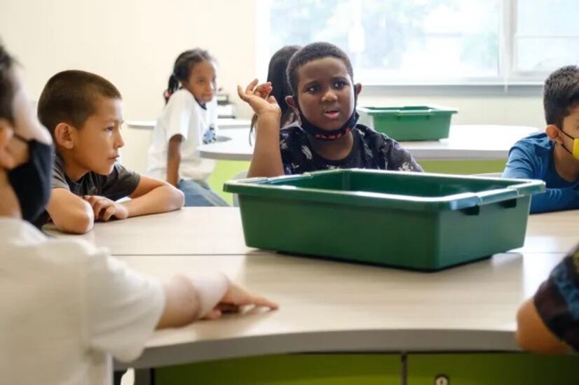Elementary students with and without masks sit around a table with a green plastic bin in the middle of the table.
