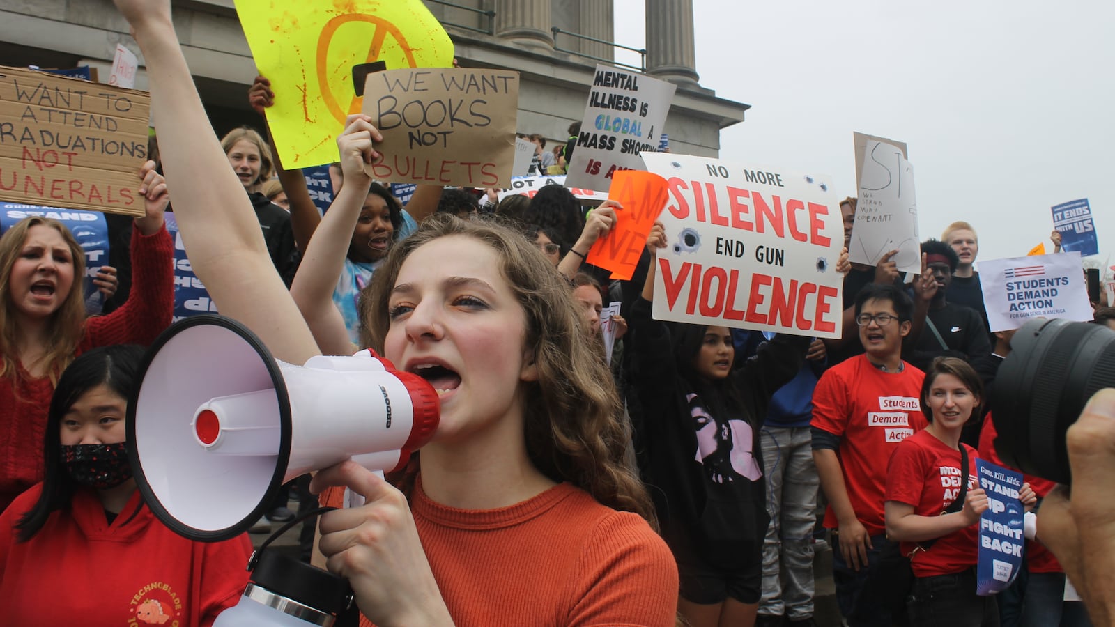 A girl yells into a megaphone surrounded by students holding signs on the steps of a large building.