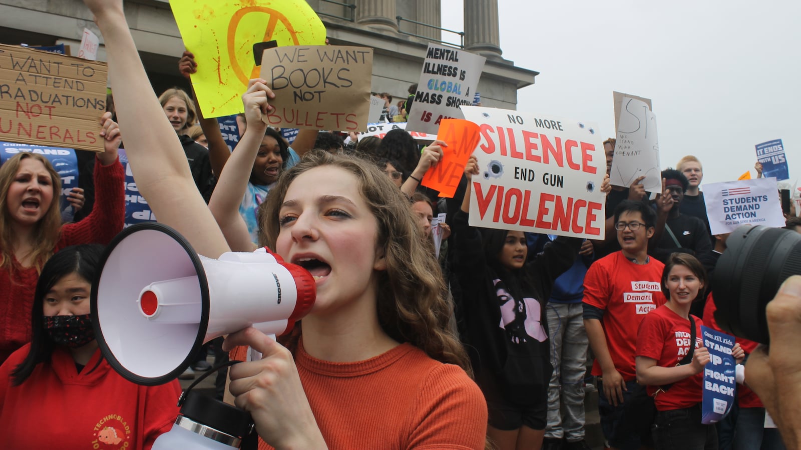 A girl yells into a megaphone surrounded by students holding signs on the steps of a large building.