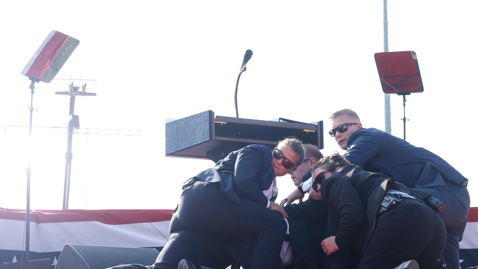 A group of Secret Service agents shields former President Donald Trump on a stage. Behind them, a podium and teleprompters.
