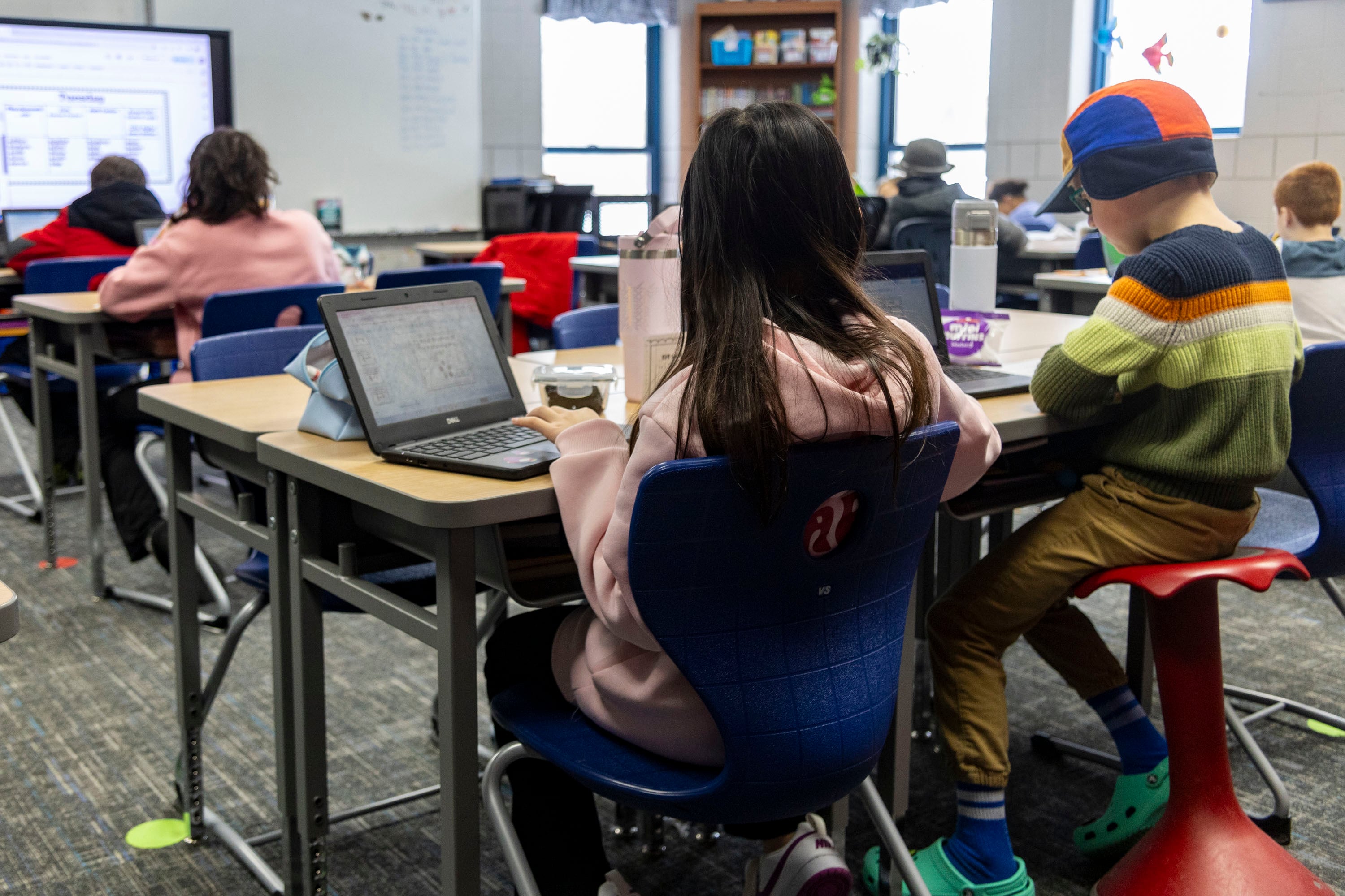 A view from behind four fourth grade students all sitting at desks in a classroom.