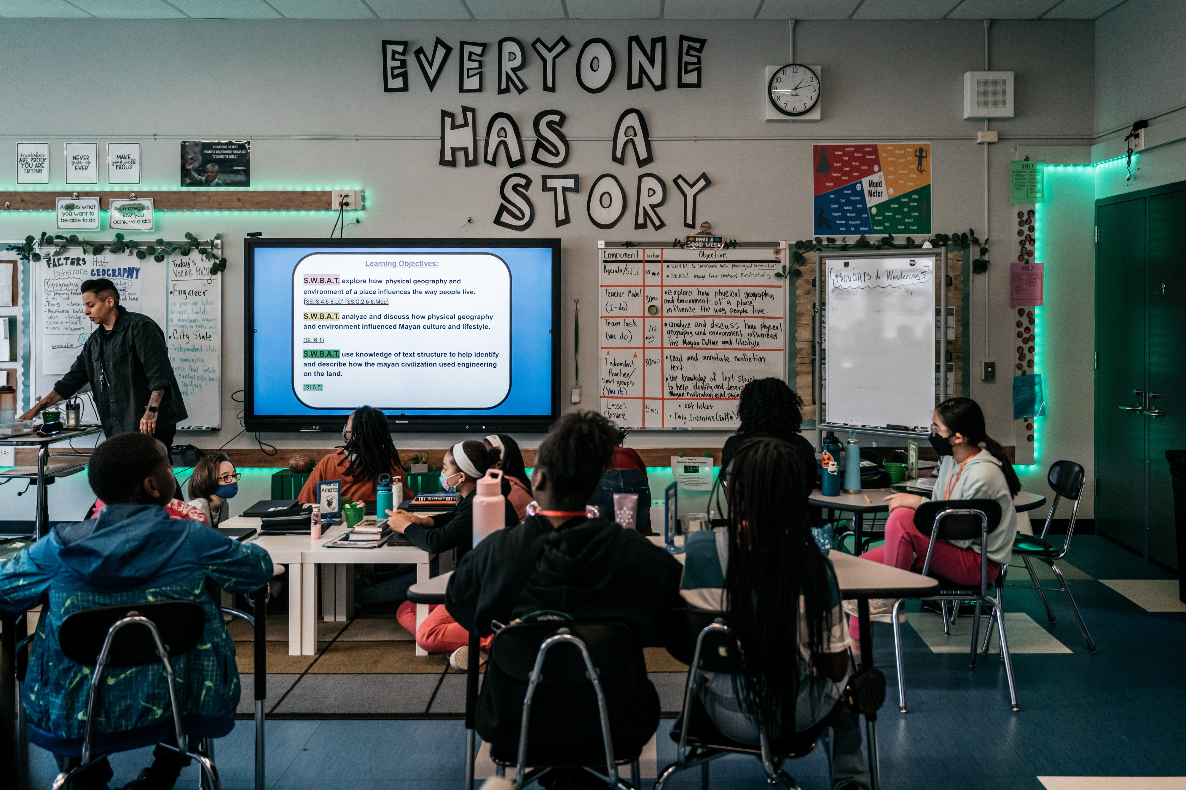 Students look up at their teacher and the lesson projection on the screen. The words “EVERYONE HAS A STORY” hang high on the classroom wall.
