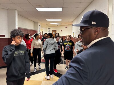 A Black man in a suit and wearing a hat faces a hallway full of middle school students.