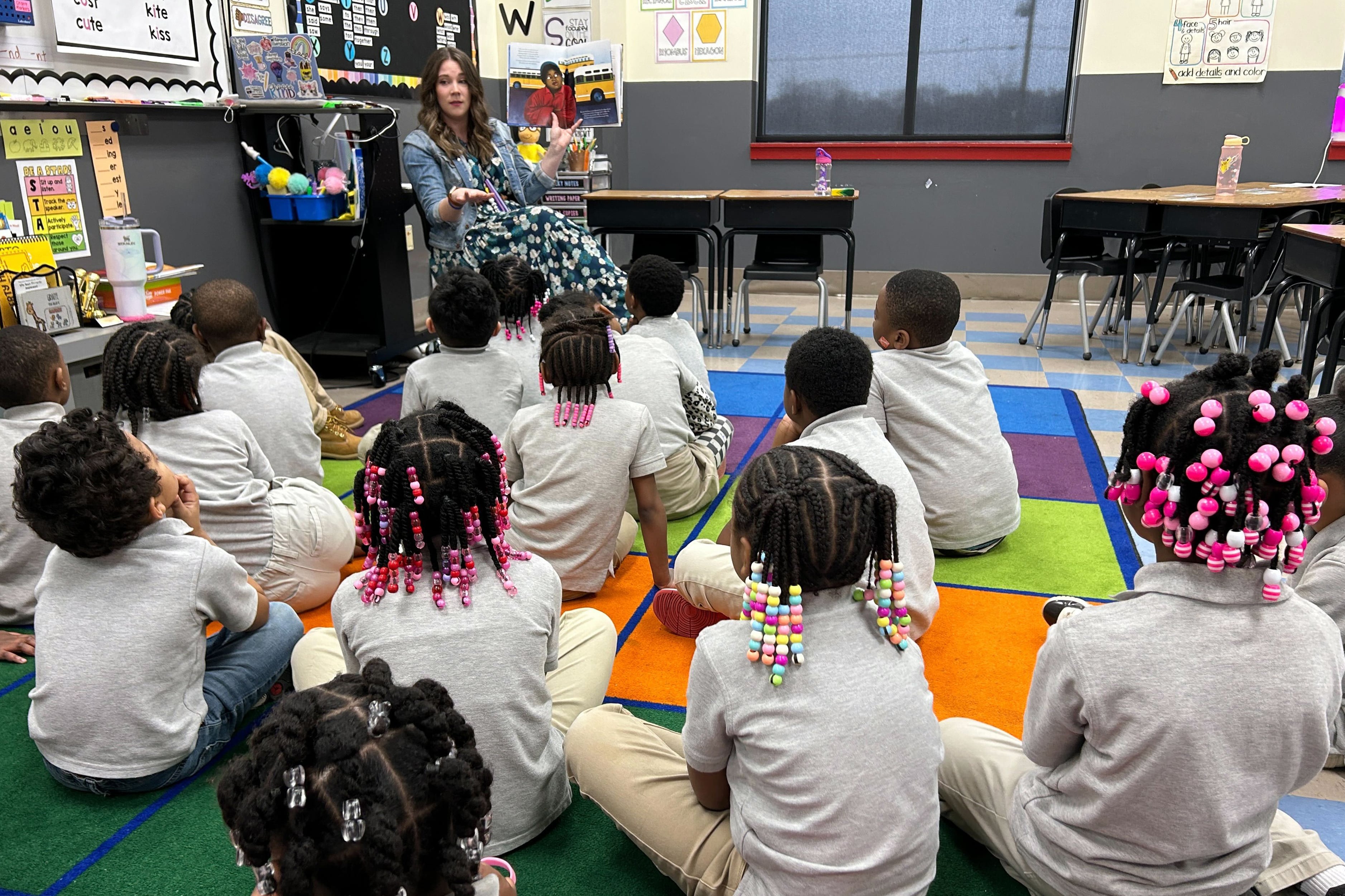A teacher with long brown hair holds up a picture book to a class full of young students sitting on a colorful rug in a classroom. Students are wearing a grey shirt and light tan pants as a uniform and many students have colorful beads in their braids.