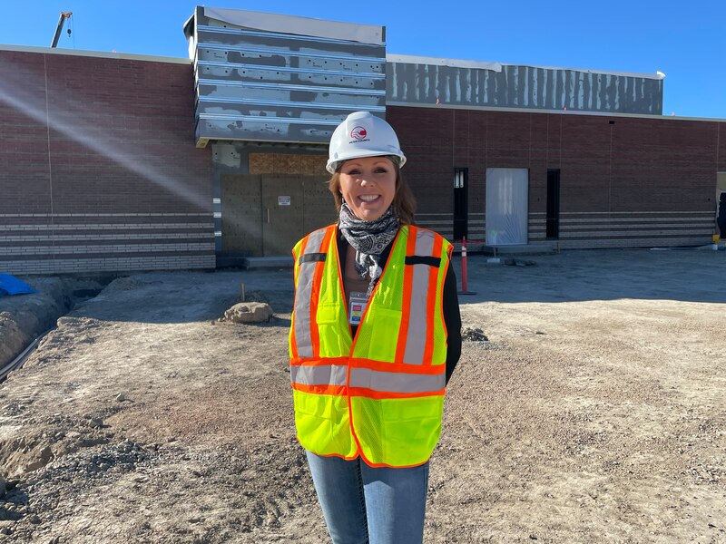 A woman wearing a safety vest and a hard hat stands in front of a building with a dirt lot in the middle ground.