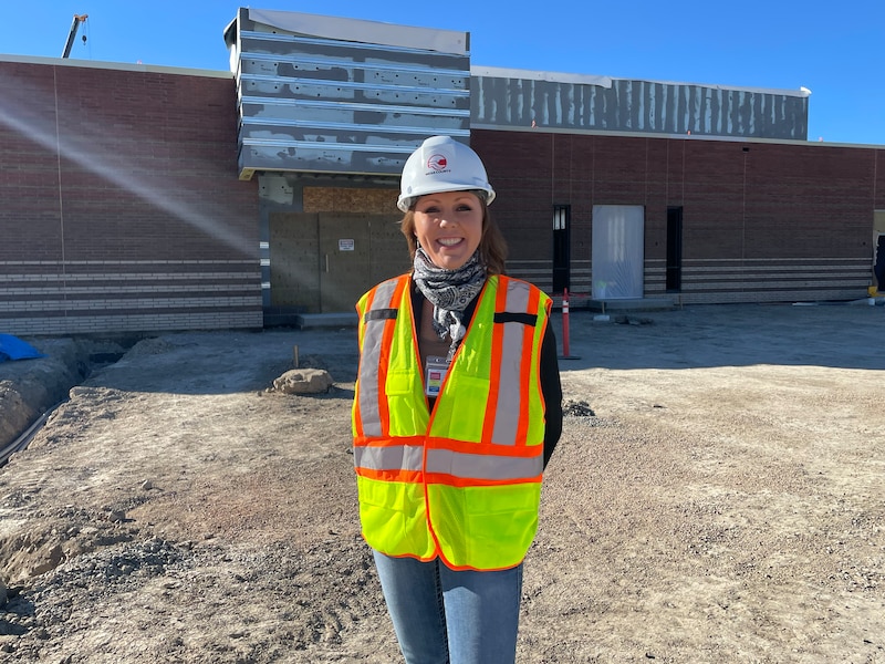 A woman wearing a safety vest and a hard hat stands in front of a building with a dirt lot in the middle ground.