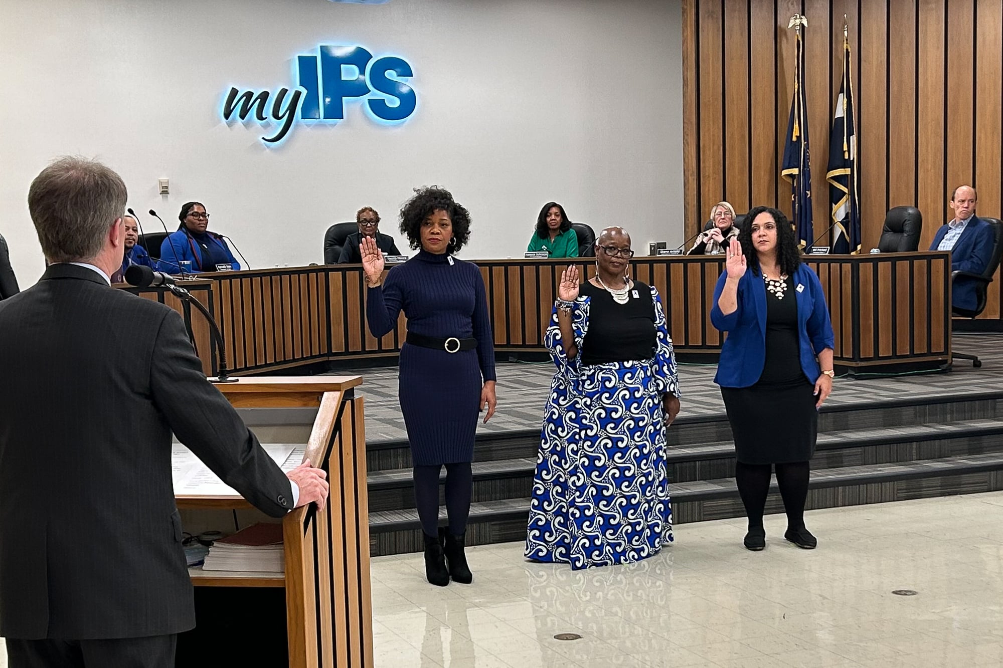 Three women stand in front of a dais and raise their right hands as people sit on the dais behind them. “myIPS” is on the wall behind them.
