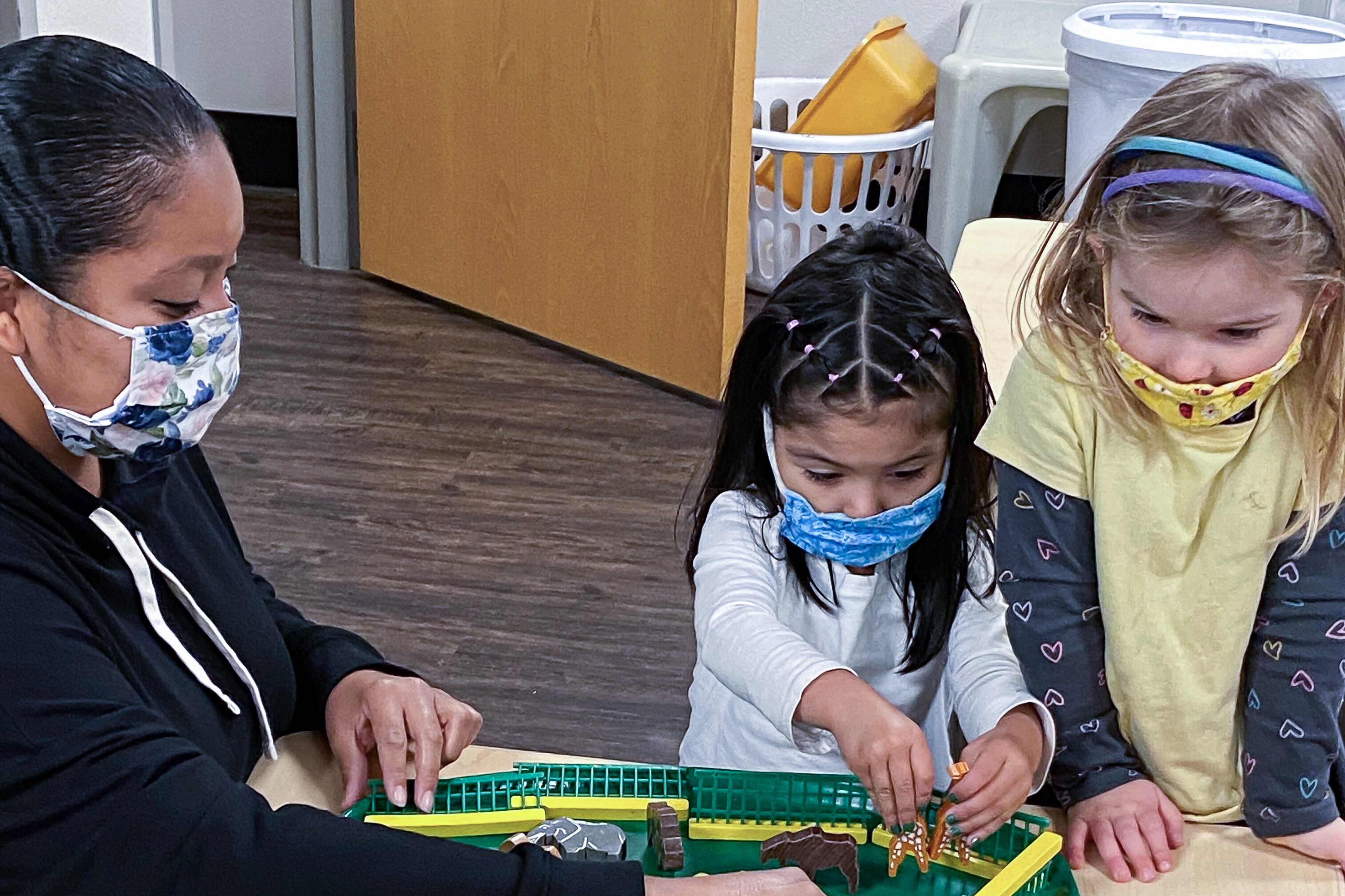 A teacher works with two preschool girls making a farm scene on a green plastic tray.