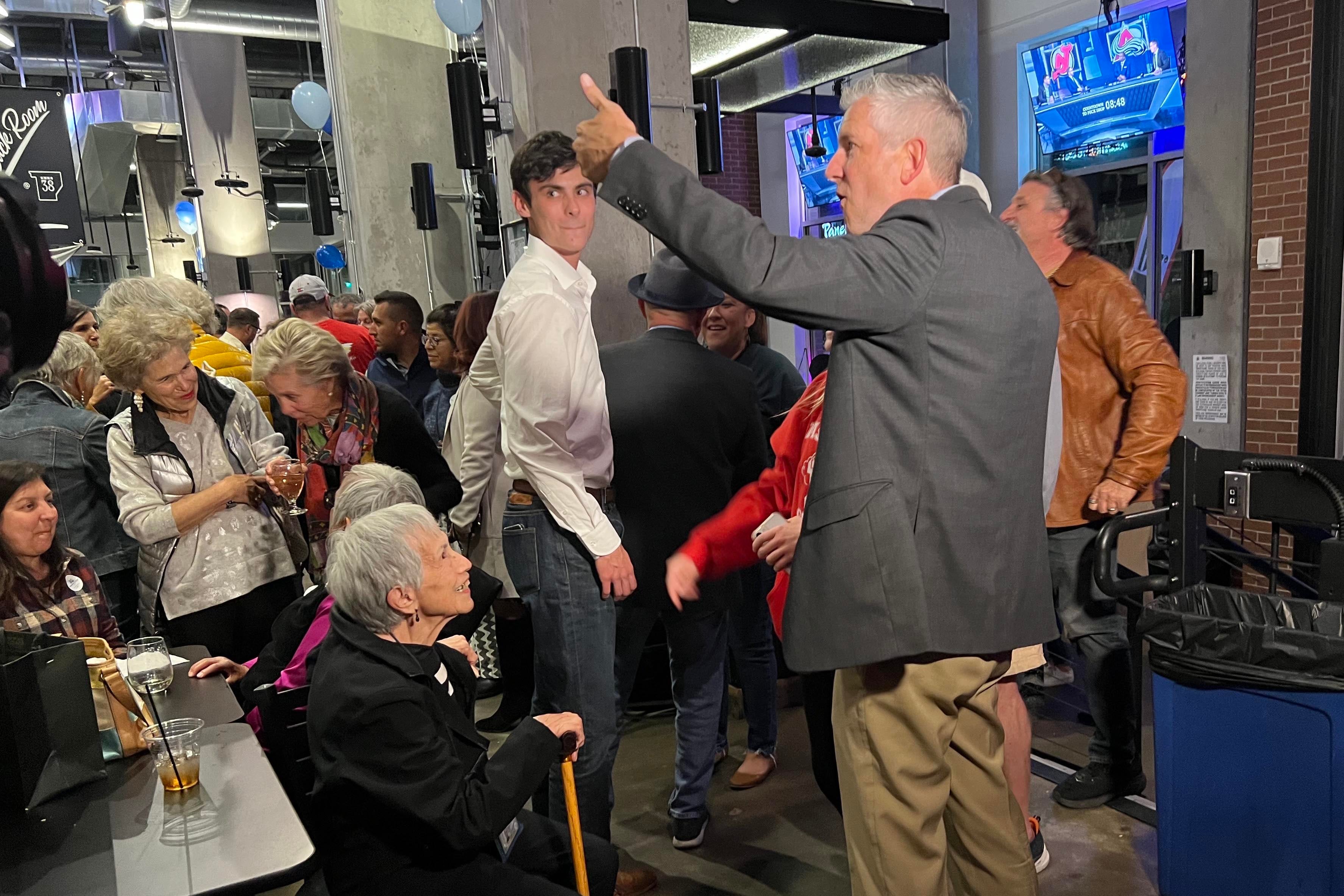 A man with white hair and a grey suit jacket with khaki pants, celebrates after winning an election in a large crowd indoors.