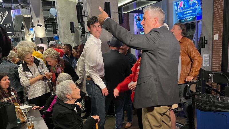 A man with white hair and a grey suit jacket with khaki pants, celebrates after winning an election in a large crowd indoors.