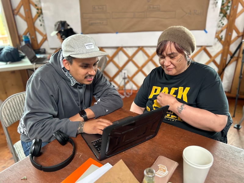 A man wearing a grey jacket and a baseball cap sits at a laptop beside a pair of headphones and a teacher wearing a T-shirt and knit cap.