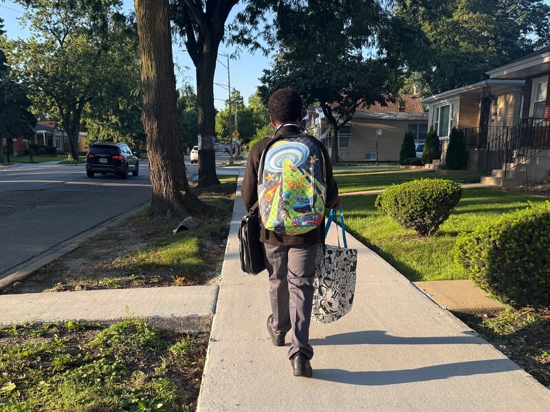 A Black young boy carrying a backpack and other bags walks down a sidewalk on a sunny morning.