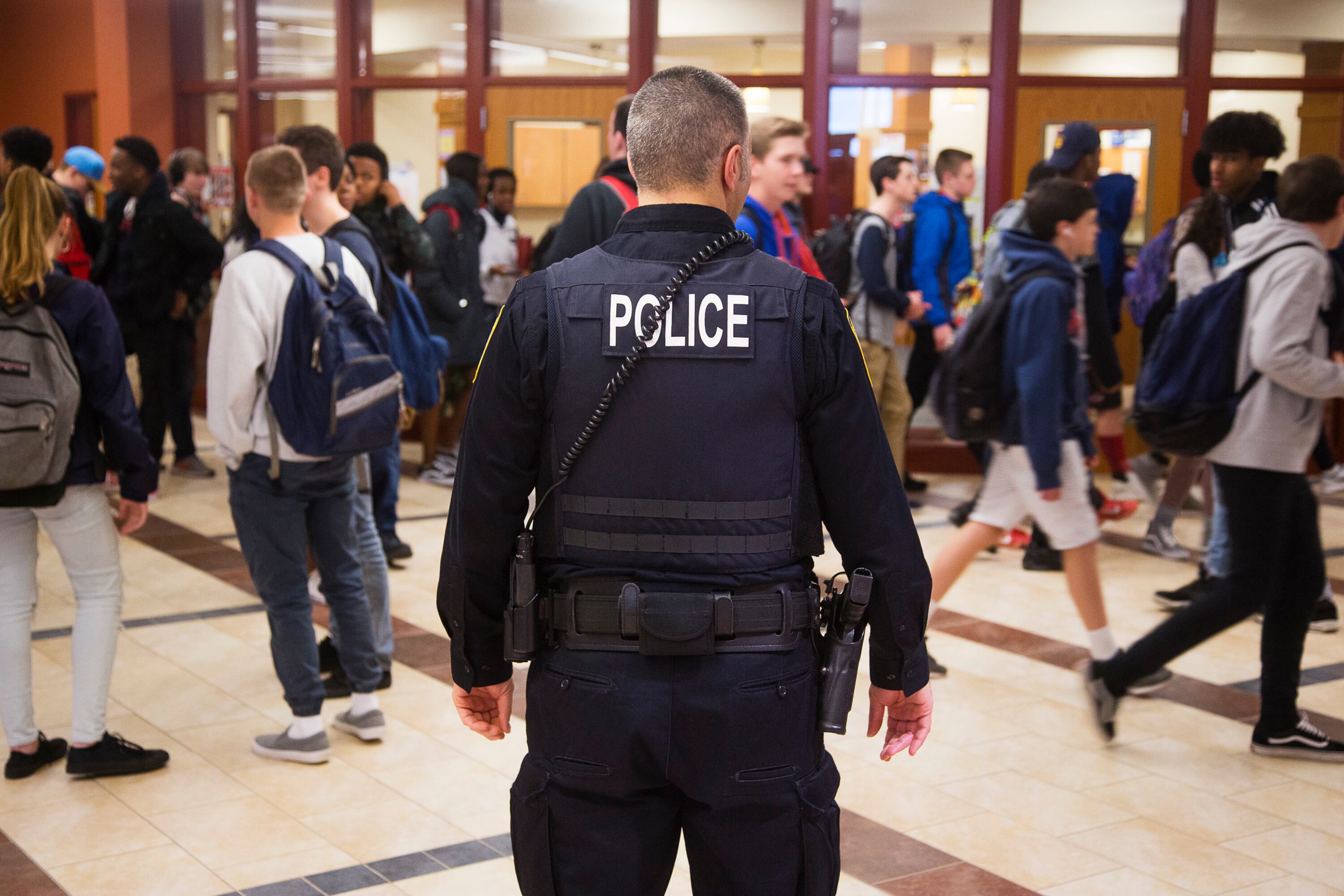 A 2018 photo shows an officer with “POLICE” marked on the back of his uniform stands in a high school hallway with students wearing backpacks walking by.
