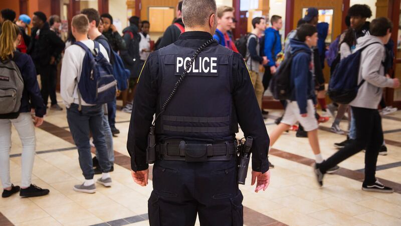 A 2018 photo shows an officer with “POLICE” marked on the back of his uniform stands in a high school hallway with students wearing backpacks walking by.