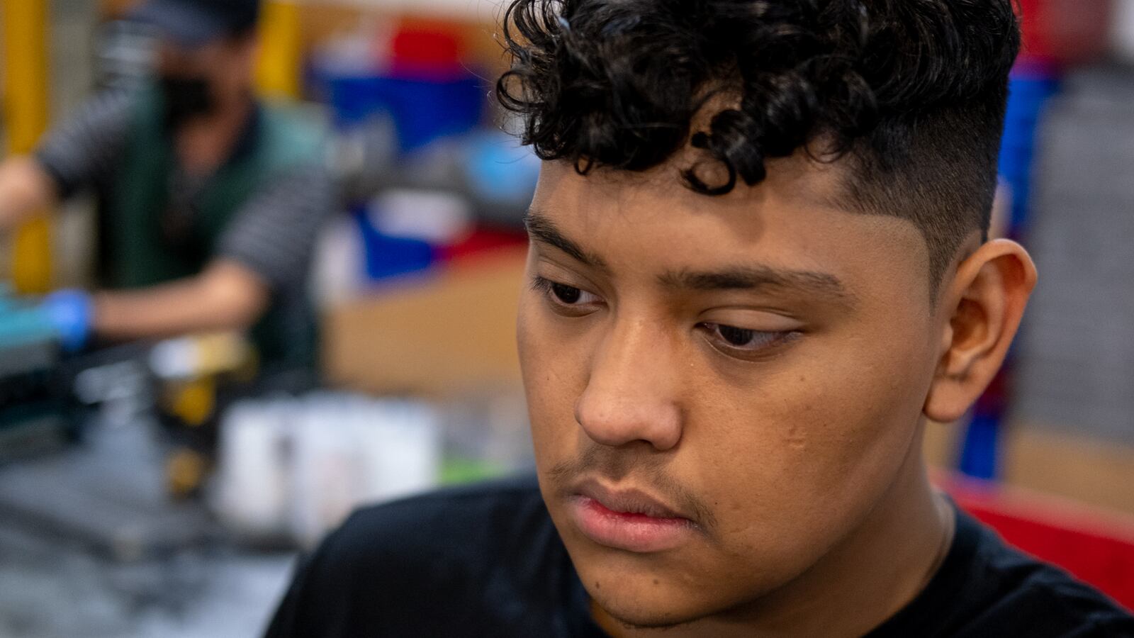 A young man wearing a black shirt with short, curled hair stares blankly while working in a factory.