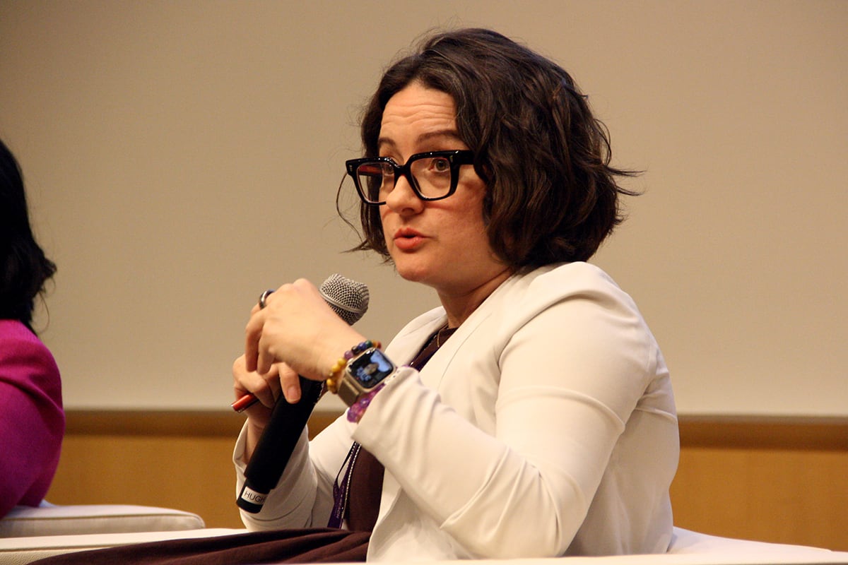 A woman with a white jacket and glasses speaks into a microphone.