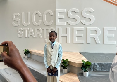A young student dressed in a nice suit vest poses for a photograph in front of a white wall that reads "Success Starts Here."