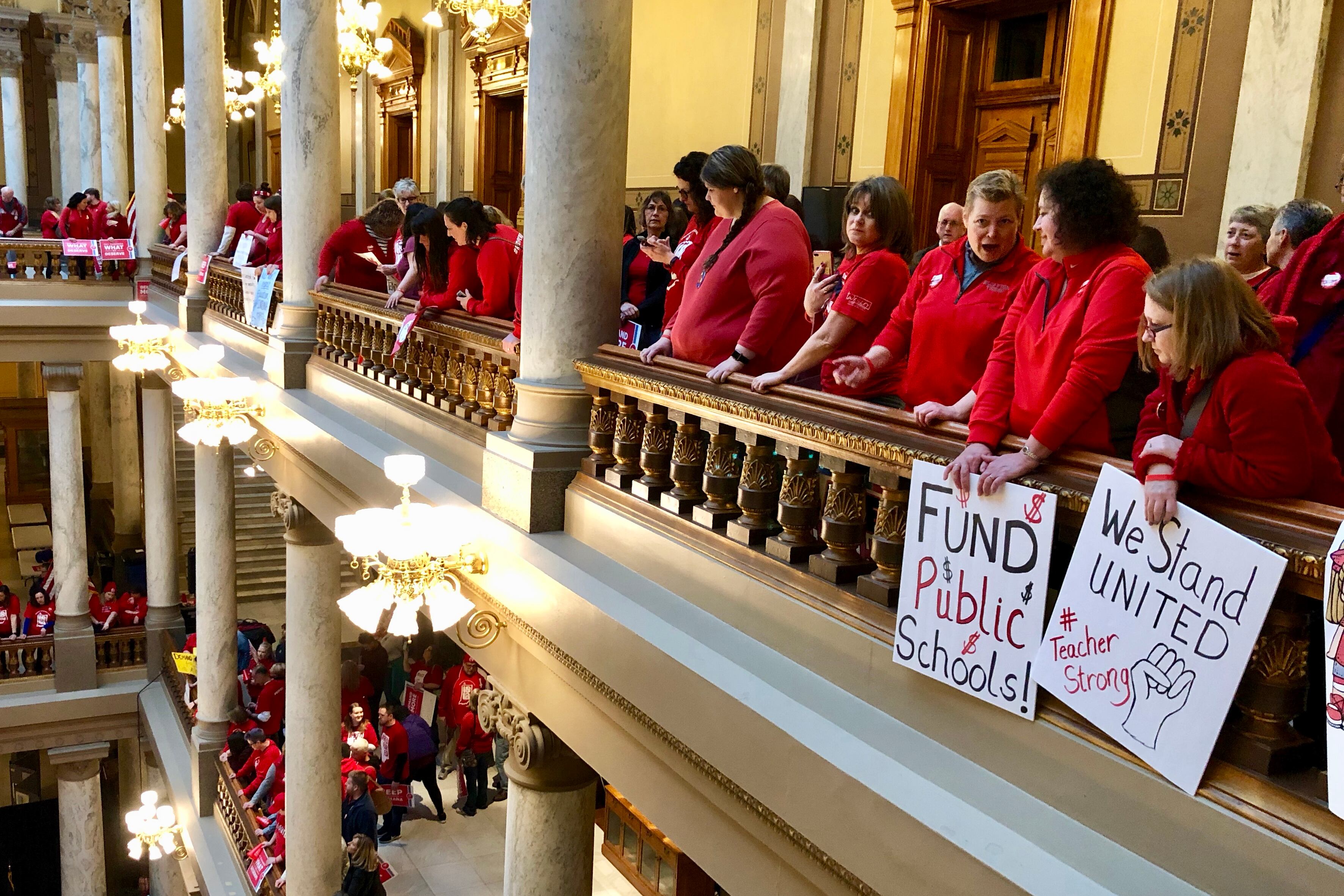 A row of teachers wearing red stand in the balcony of the state Capitol on March 9, 2019, at a rally hosted by the Indiana State Teachers Association. Two hold placards saying “Fund Public Schools” and “We Stand United.”