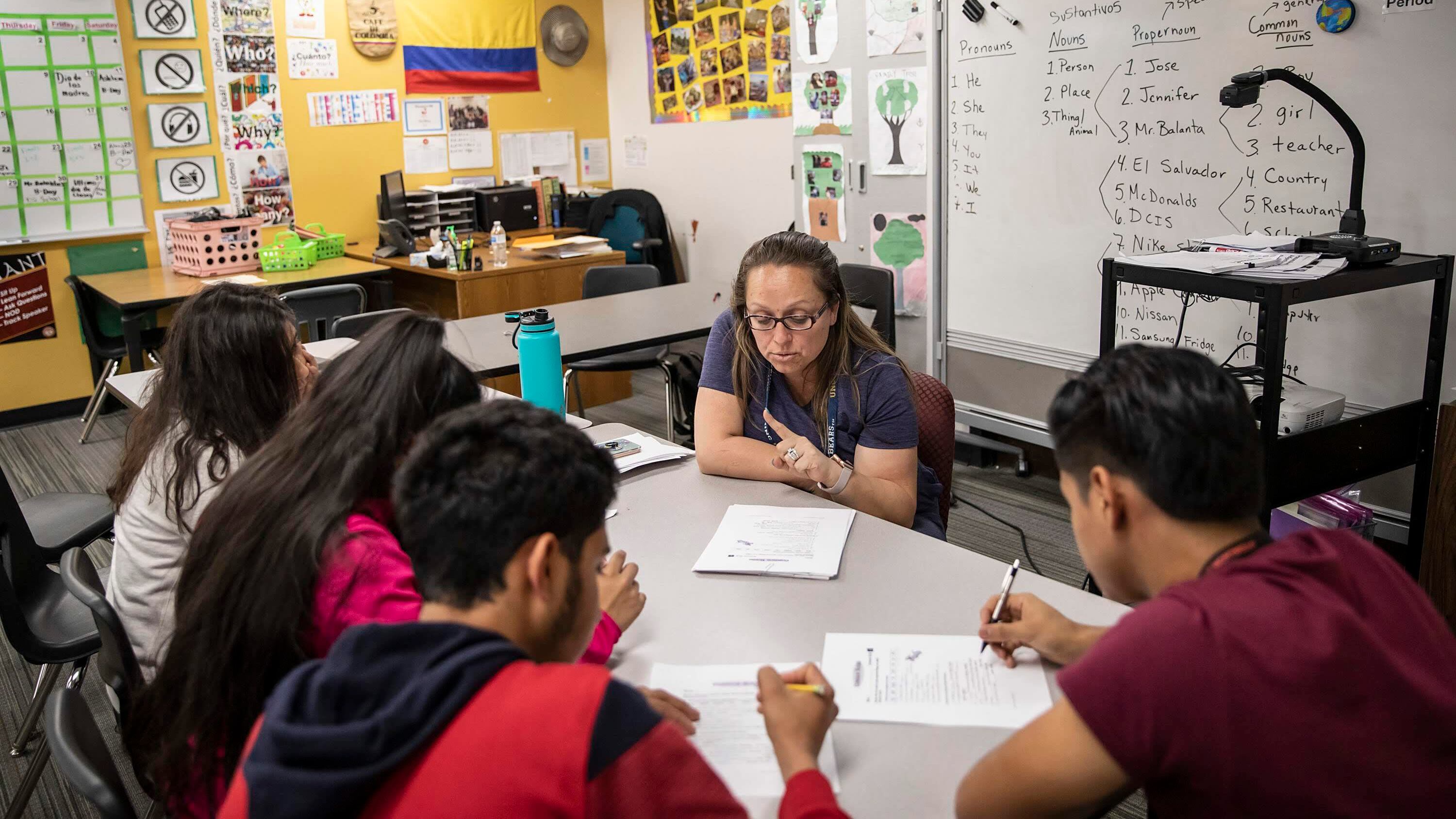 A woman wearing glasses and a school lanyard sits at a rectangular table with four adolescent students as she holds her finger up while explaining. The walls of the classroom are covered in posters and signs with English keywords and vocabulary.