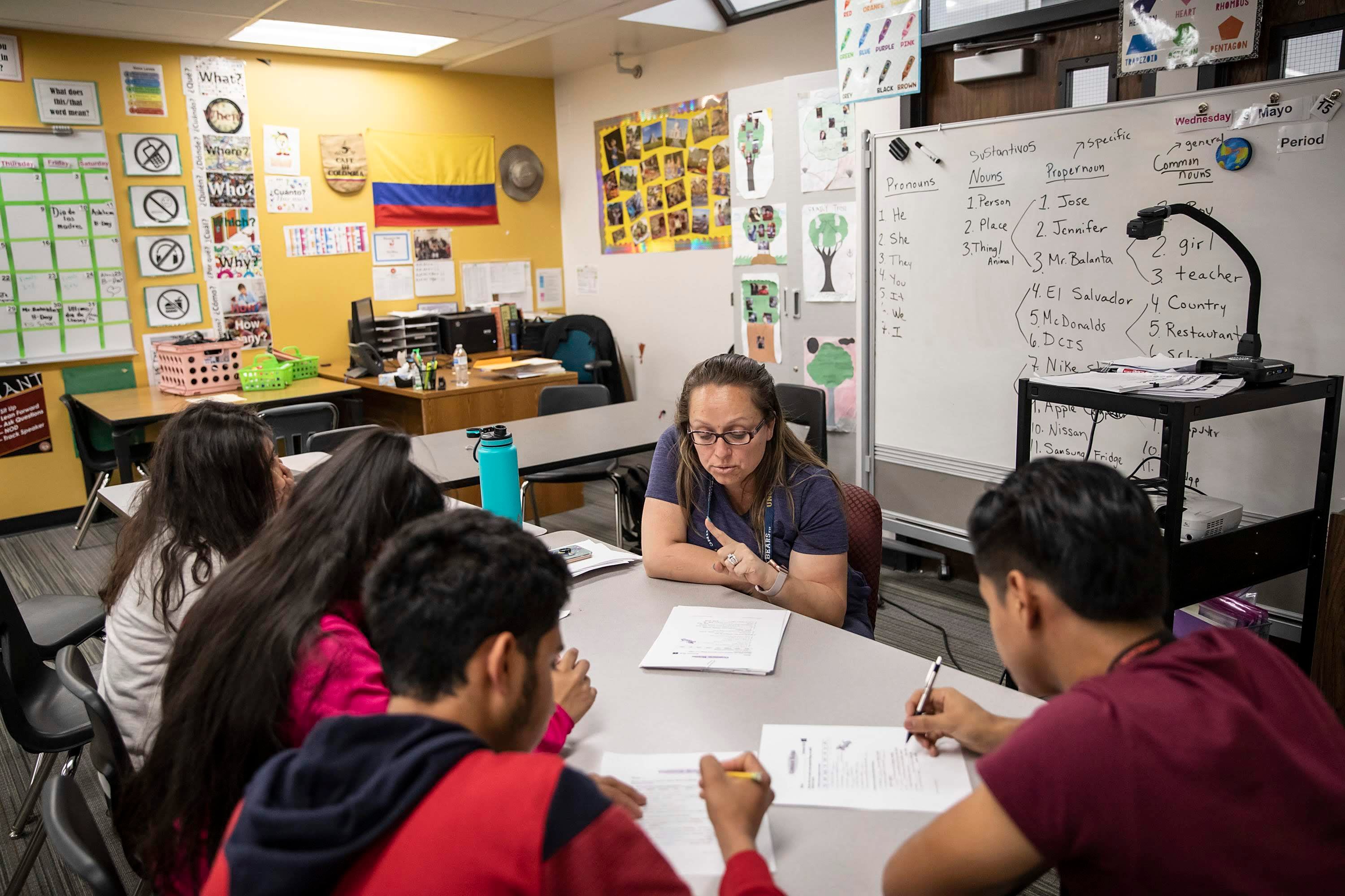 A woman wearing glasses and a school lanyard sits at a rectangular table with four adolescent students as she holds her finger up while explaining. The walls of the classroom are covered in posters and signs with English keywords and vocabulary.