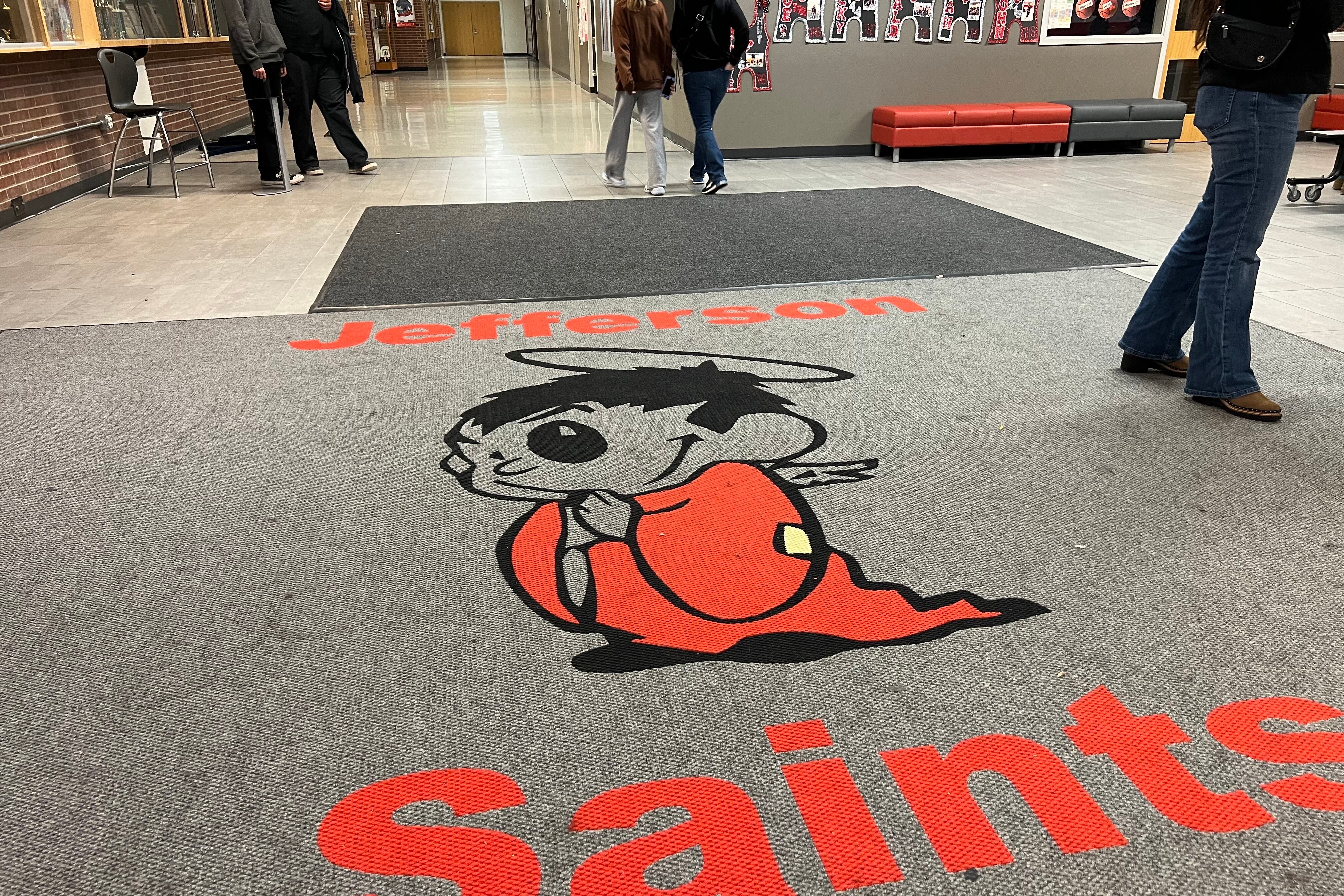 A high school foyer with a giant gray rug that says "Jefferson Saints" and has a cartoon of a red saint mascot.