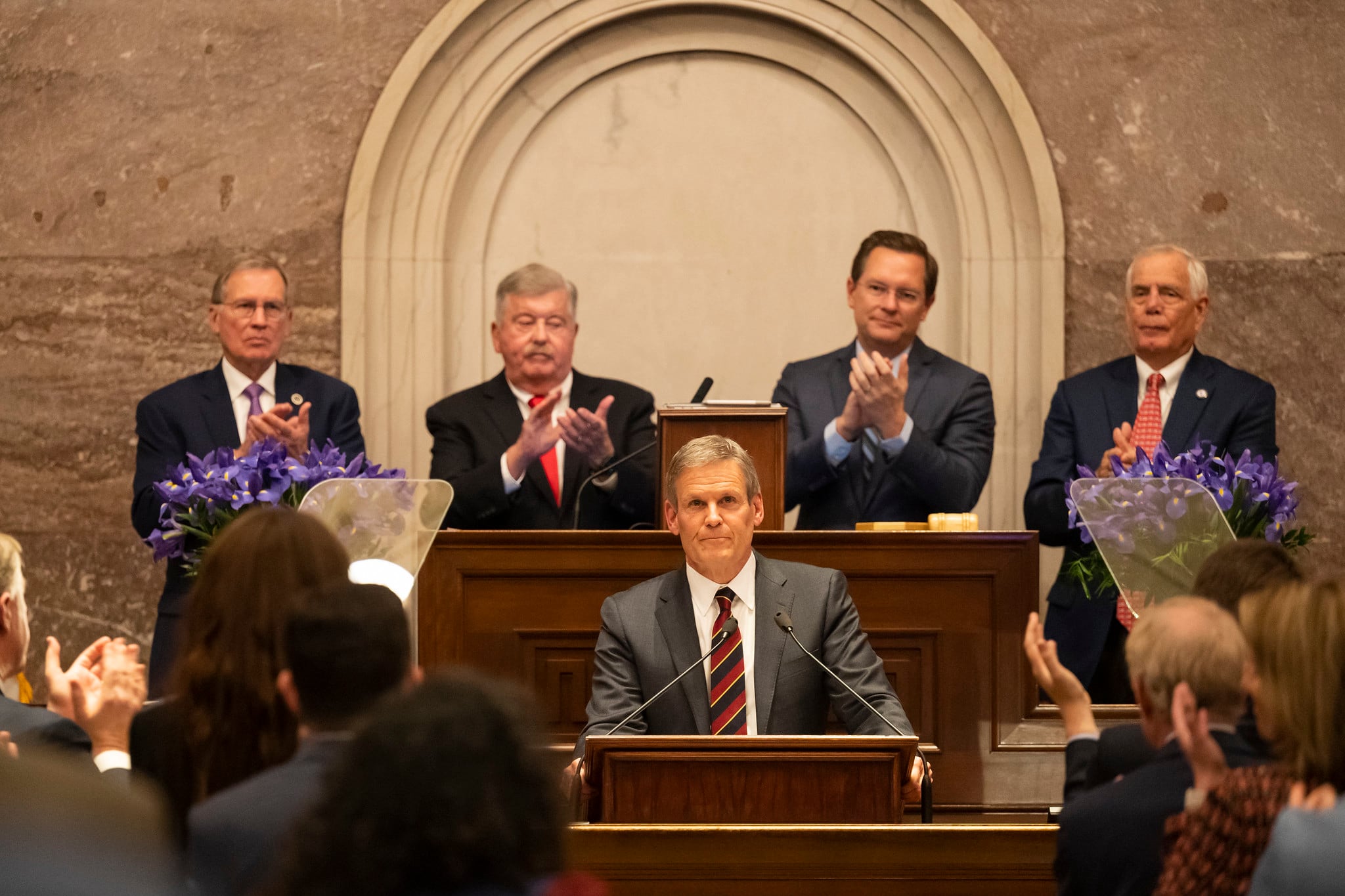 A man in a business suit stands at a podium surrounded by other men who are clapping.