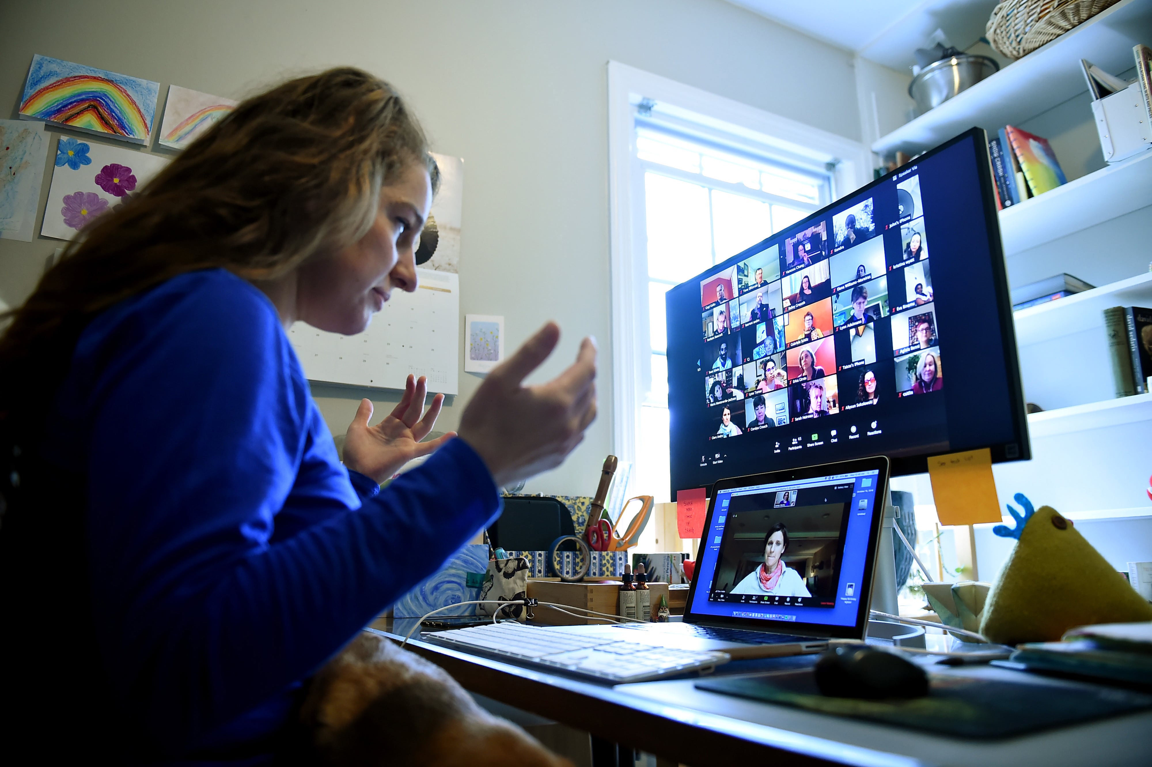 A teacher seated at a desk looks into a laptop and screen during remote learning.