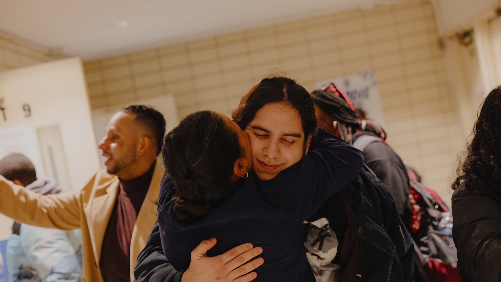 A photograph of an adult woman embracing an older high school student in a crowded hallway. There is a man on the left of them holding a cellphone that isn't visible in the photo to make a video.