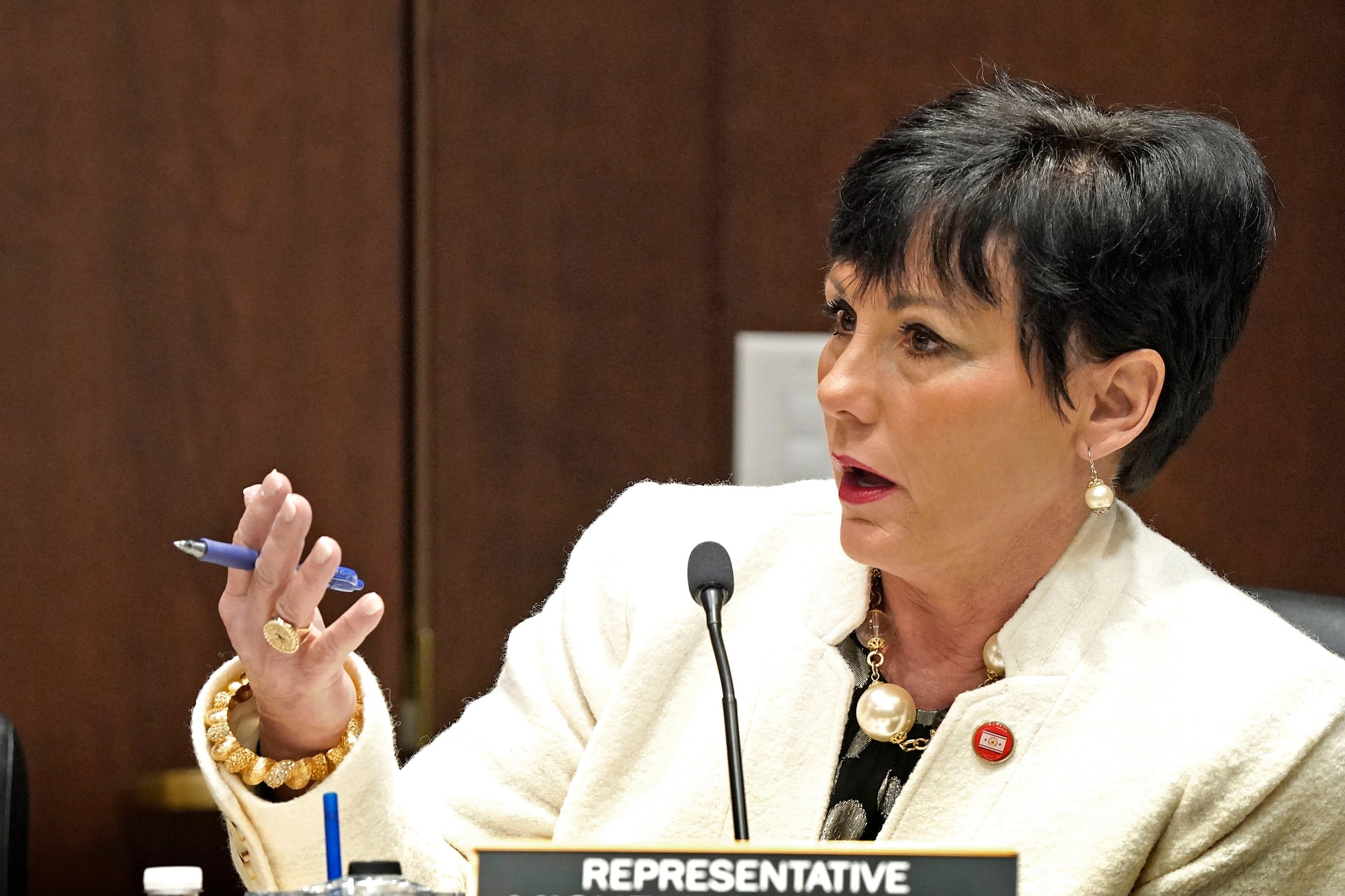 A woman in a white business suit gestures as she speaks into a microphone in a legislative chamber.