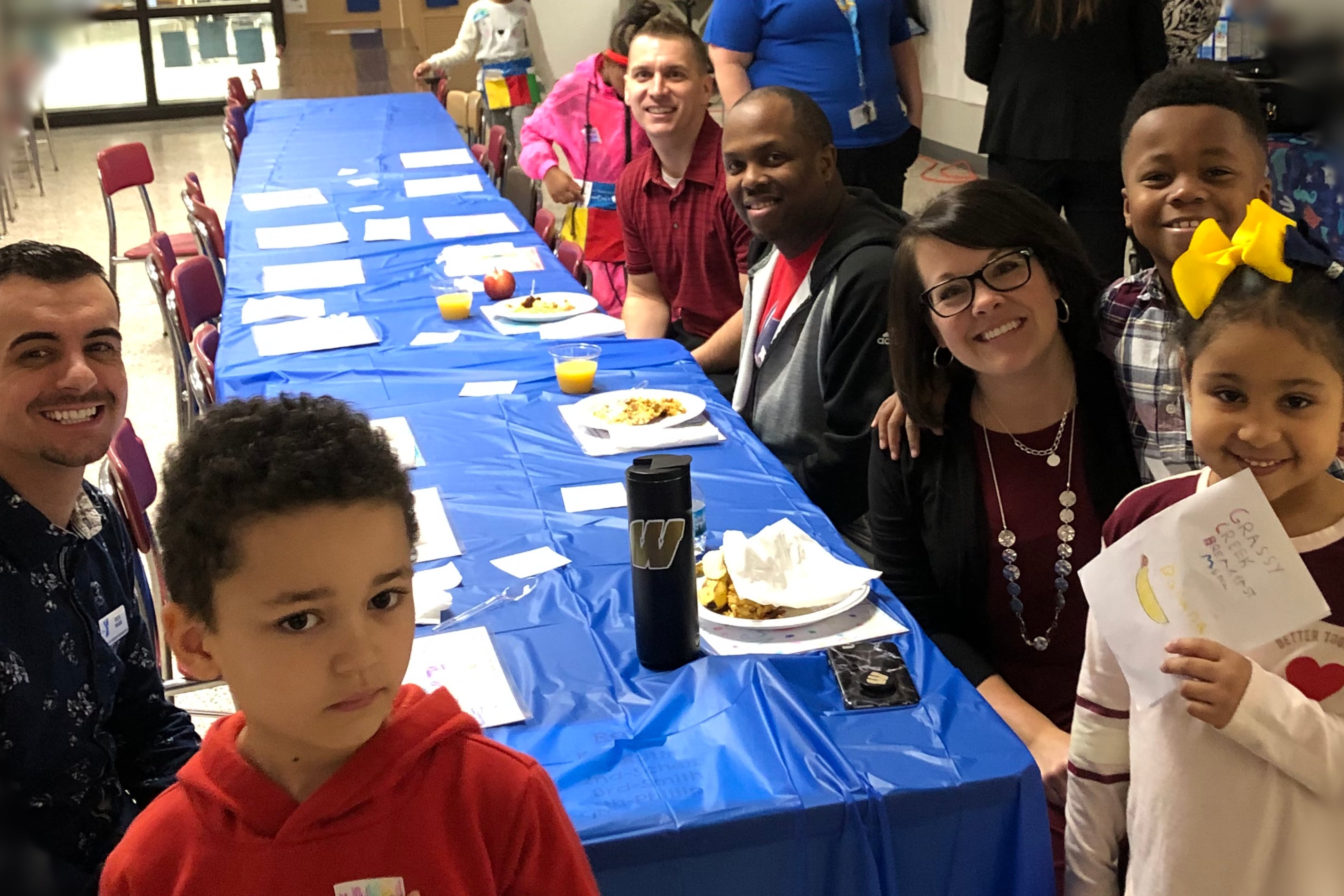 Grassy Creek Elementary School Principal Christy Merchant, students, and staff smile while sitting at a long blue table for breakfast.