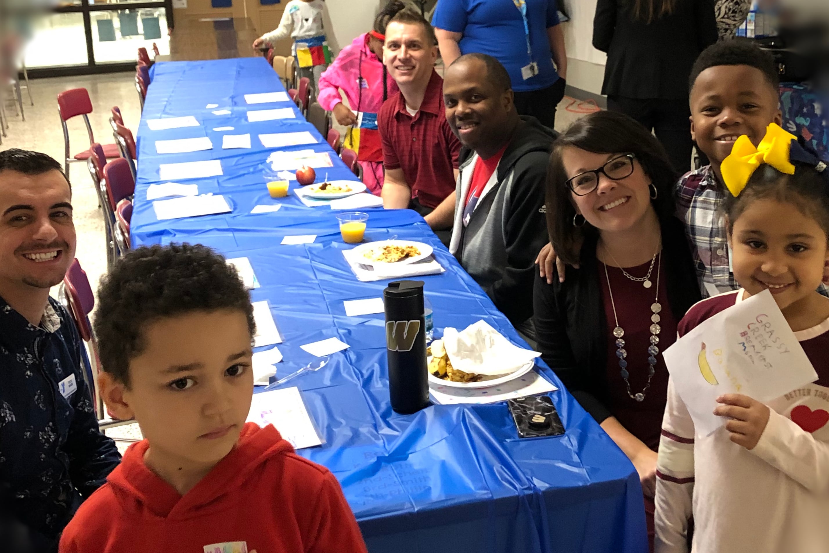 Grassy Creek Elementary School Principal Christy Merchant, students, and staff smile while sitting at a long blue table for breakfast.