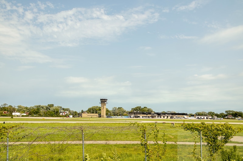An airport is seen from far away with blue sky and clouds above and green grassy field in the foreground.
