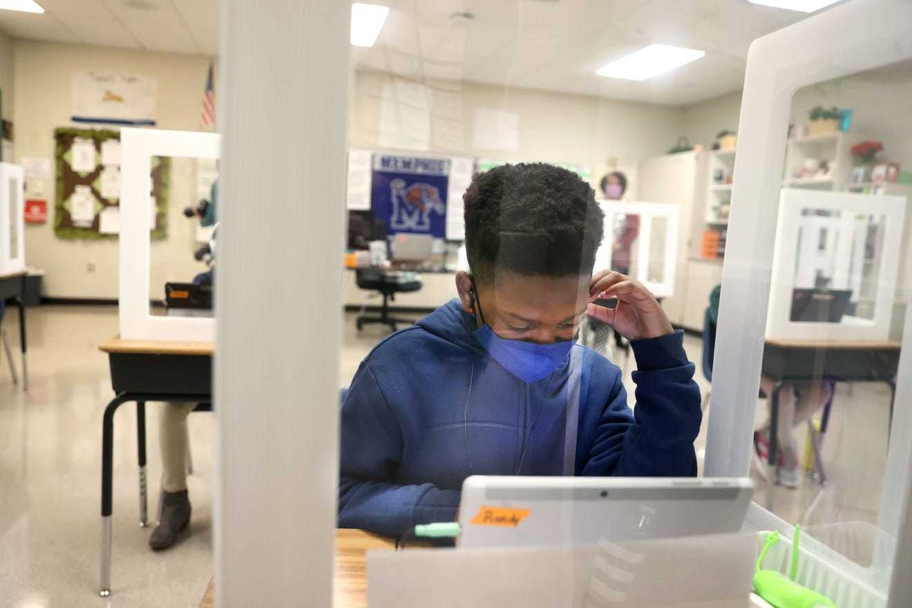A boy wearing a blue sweatshirt and blue mask sitting behind a glass partition at his desk works on his computer.