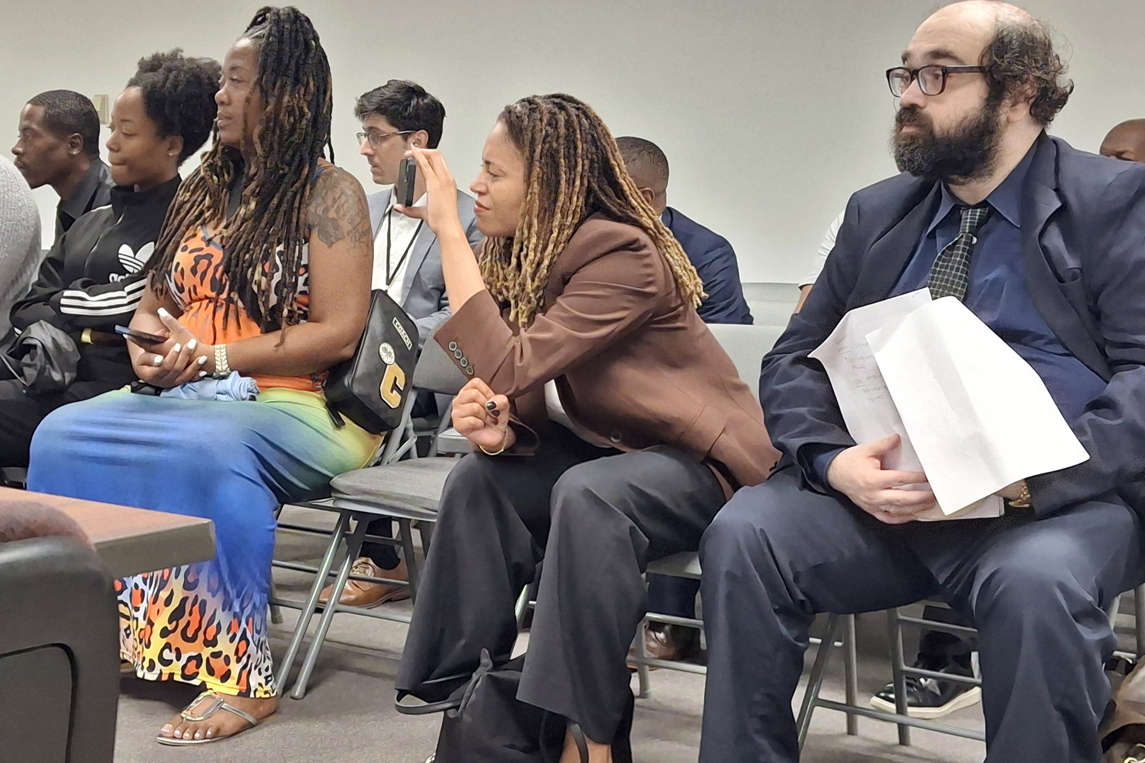 A group of adults sit in chairs facing the same way, while one Black woman with long hair holds a cellphone recording a meeting.
