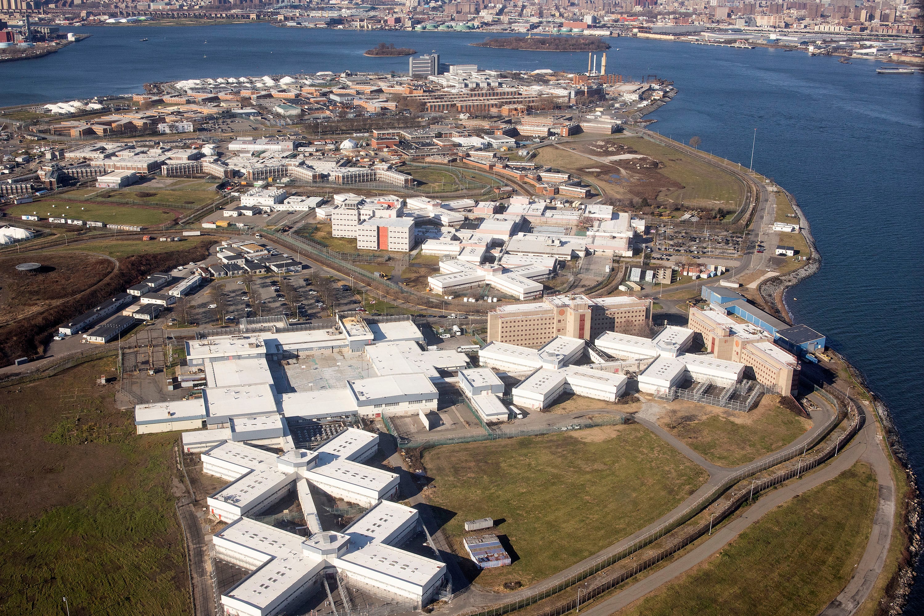 A bird's eye view of part of an island with a prison facility taking up most of the space with a river surrounding the island and a cityscape in the background.