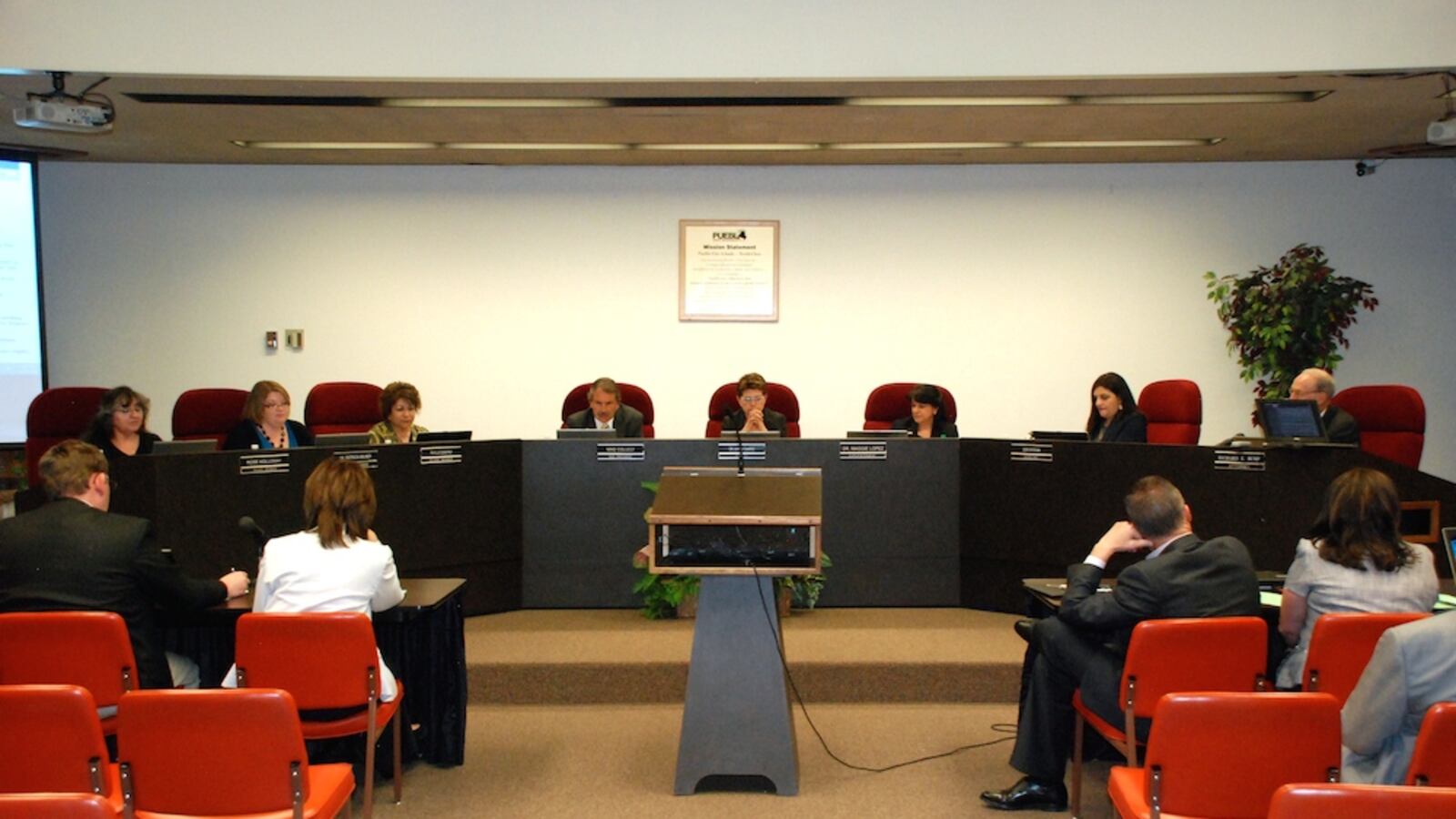 Eight people sit on a dais behind a panel, with about six people in orange chairs in the audience.