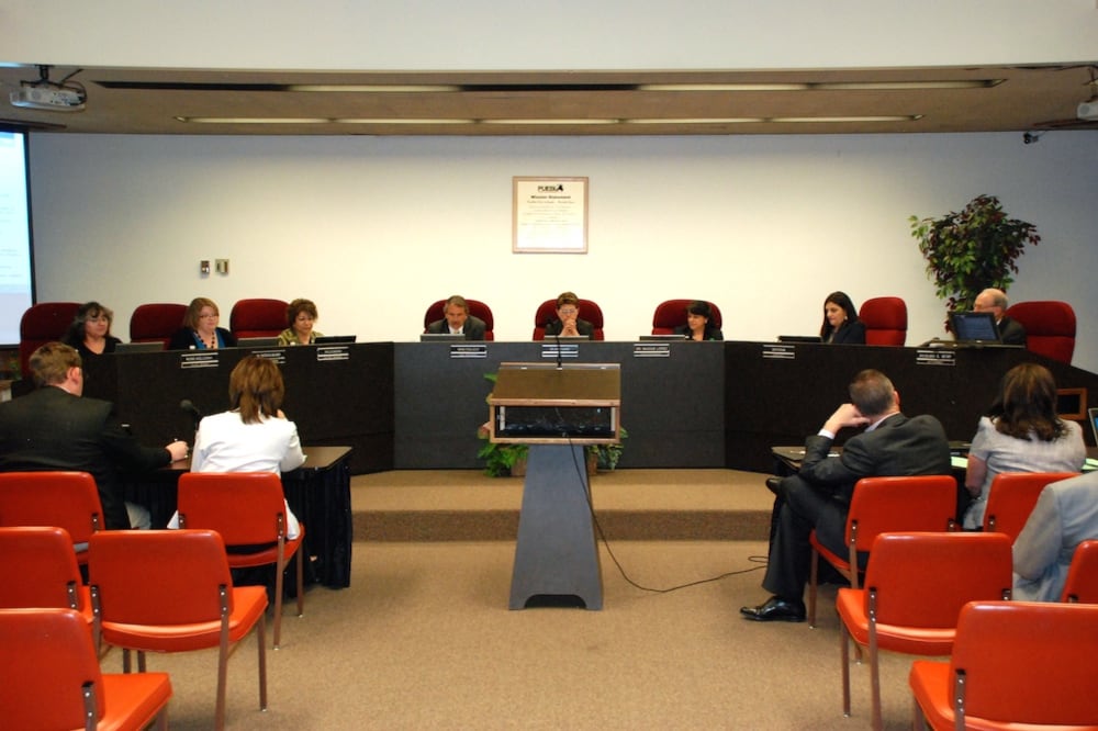 Eight people sit on a dais behind a panel, with about six people in orange chairs in the audience.
