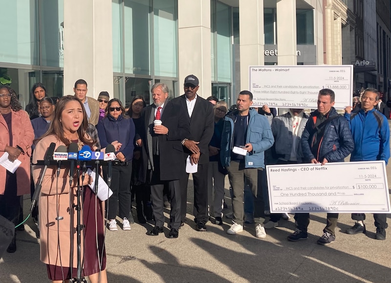 A woman speaks at a podium while a group of people looks on in a street.