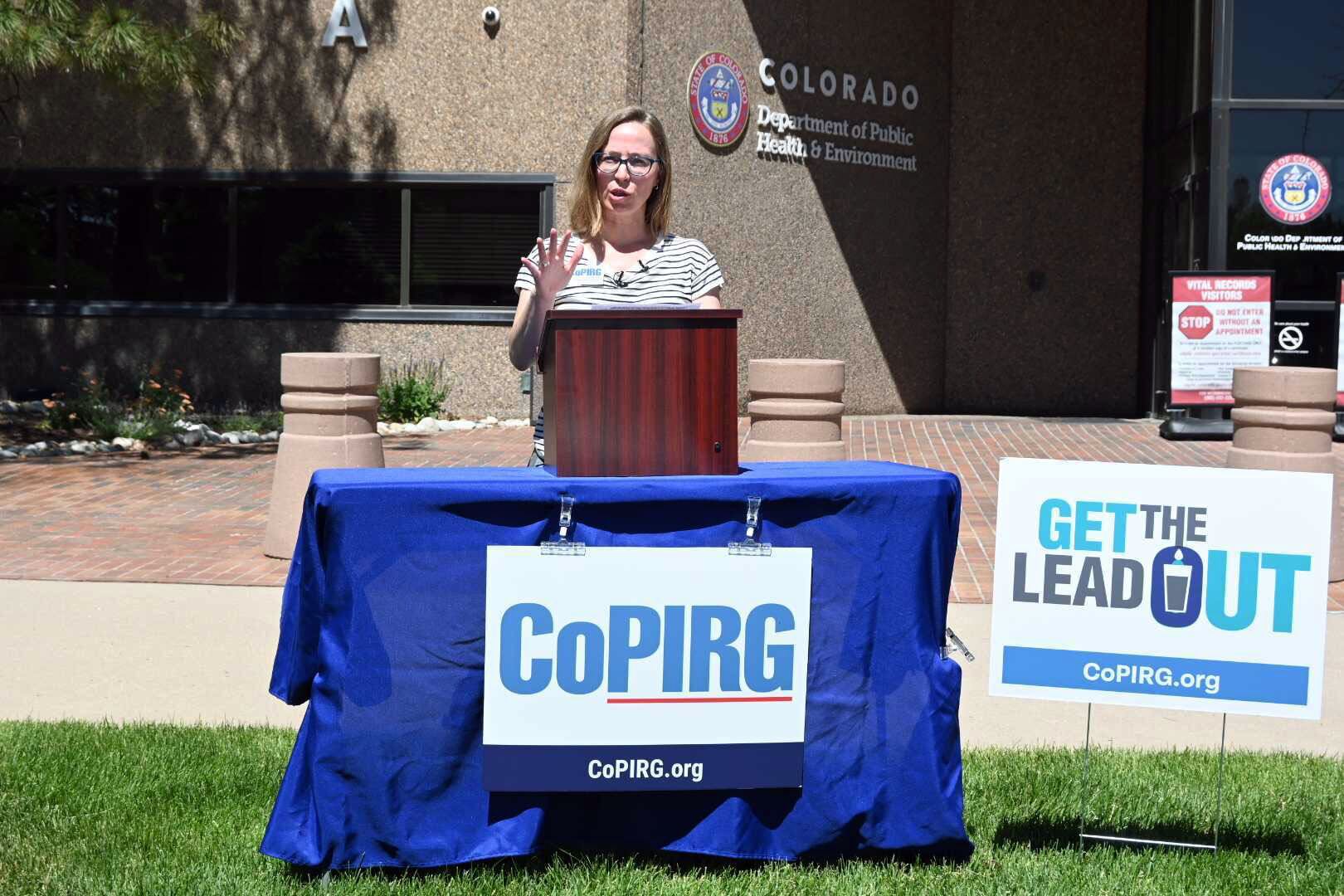 An adult woman with blonde hair and wearing glasses speaks from behind a wooden podium and a table with a blue cloth with a sign that reads "CoPRIG" outside of a large stone government building.