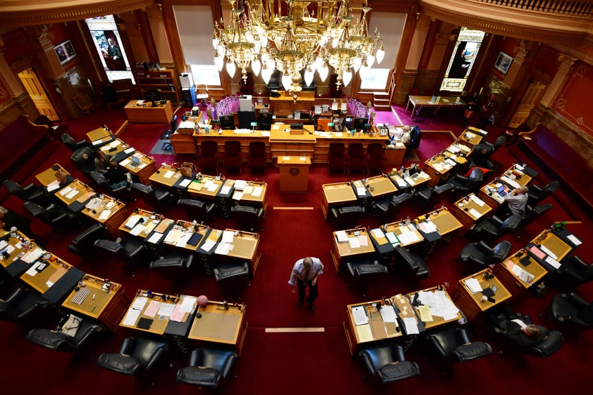 A lone senator walks down the center aisle of the Colorado State Senate. Desks are arrayed in a semi-circle facing an elaborate dais.