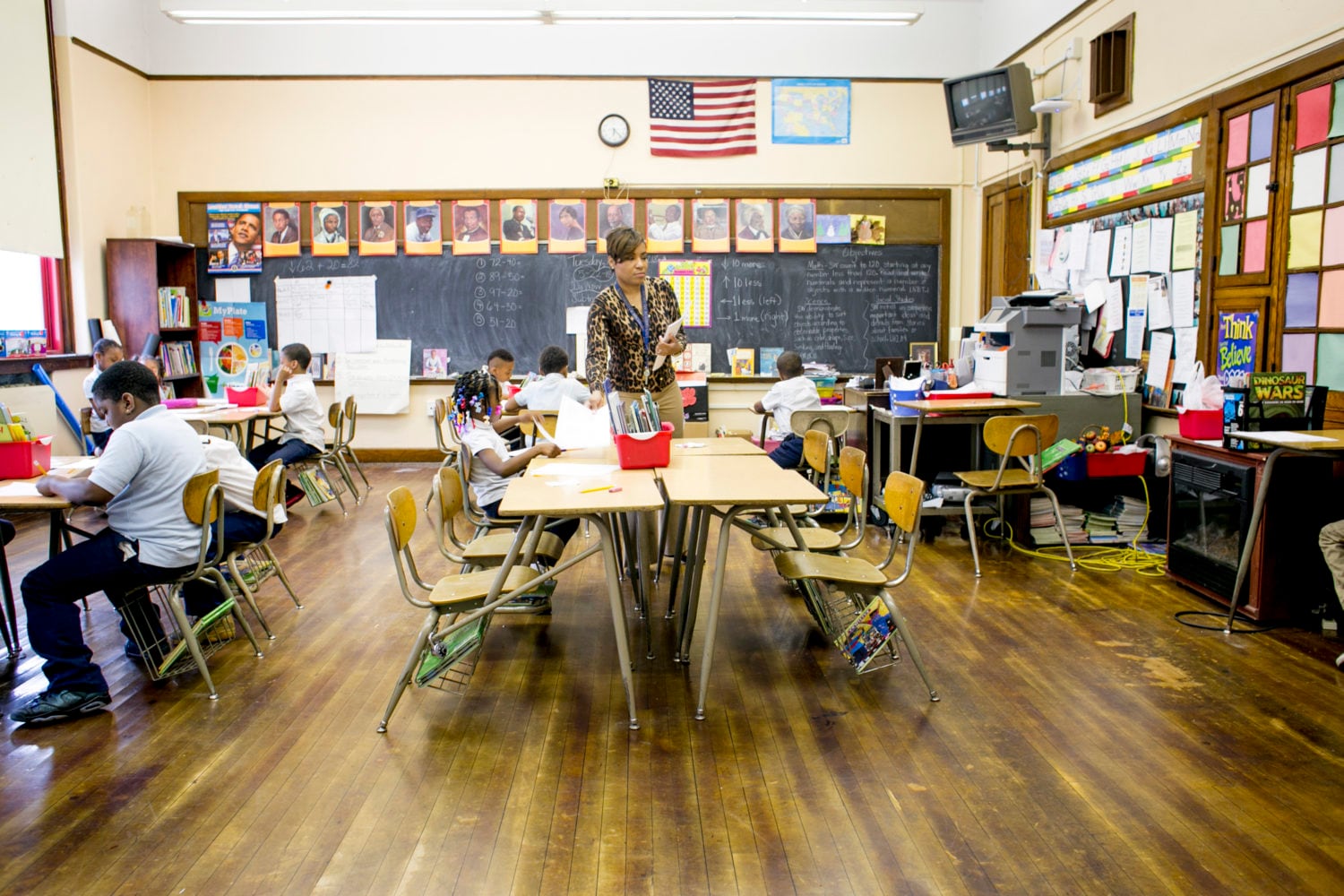 A teacher stands passes along a paper sheet to a first grade student sitting at her desk in the middle of a classroom. The classroom walls are lined with photos of prominent African and African American figures. Students are busy working on class assignments.