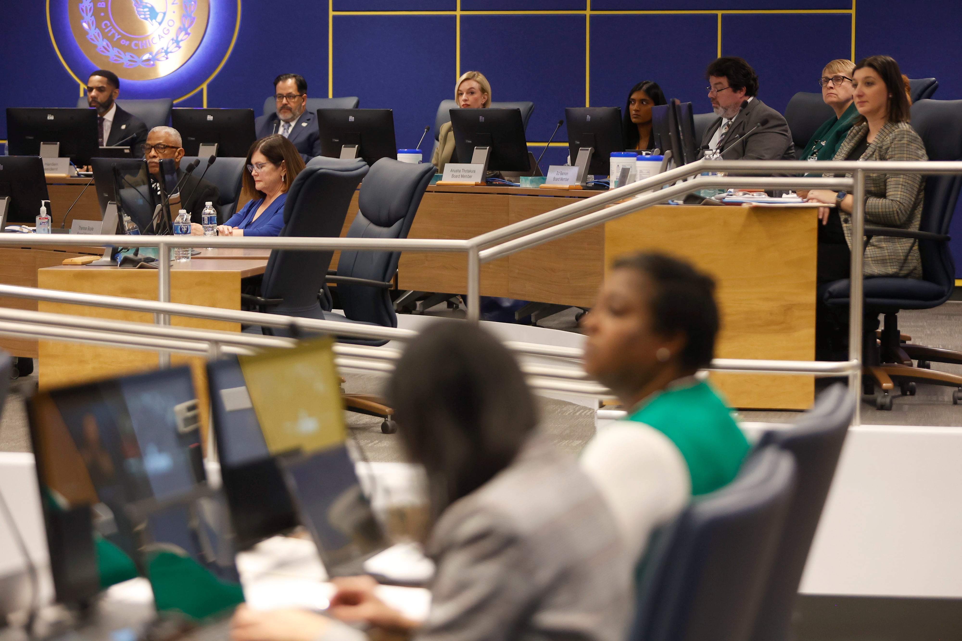 People in business clothes sit in a large conference room with a blue wall and an illuminated circular sign in the background.
