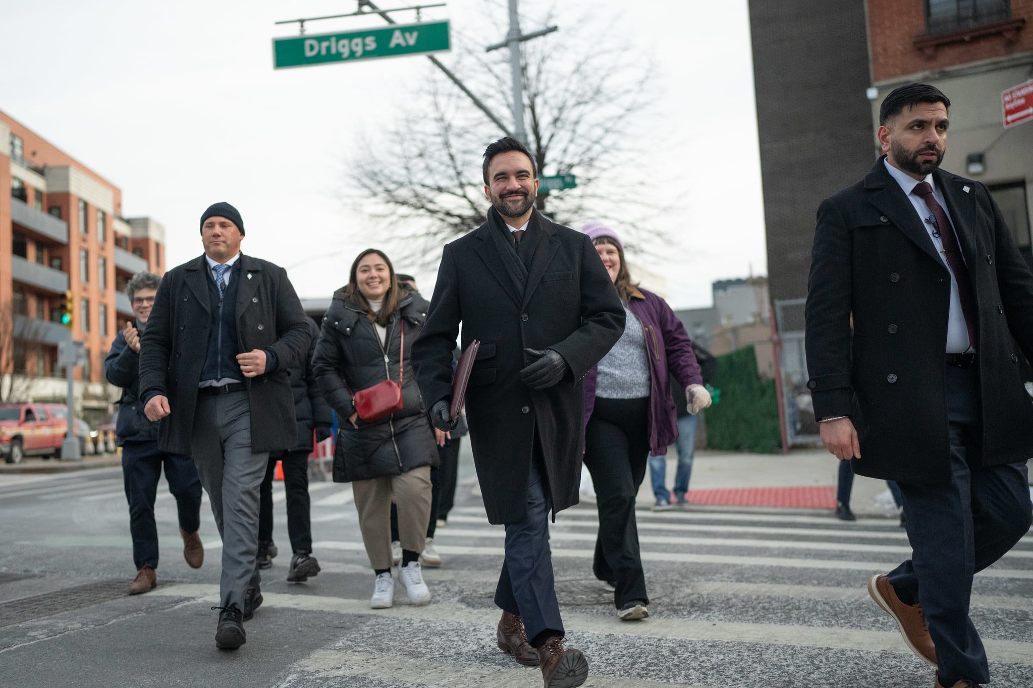 A photograph of a group of adult in suits and jackets cross the street at a cross walk on a cloudy, cold winter day.