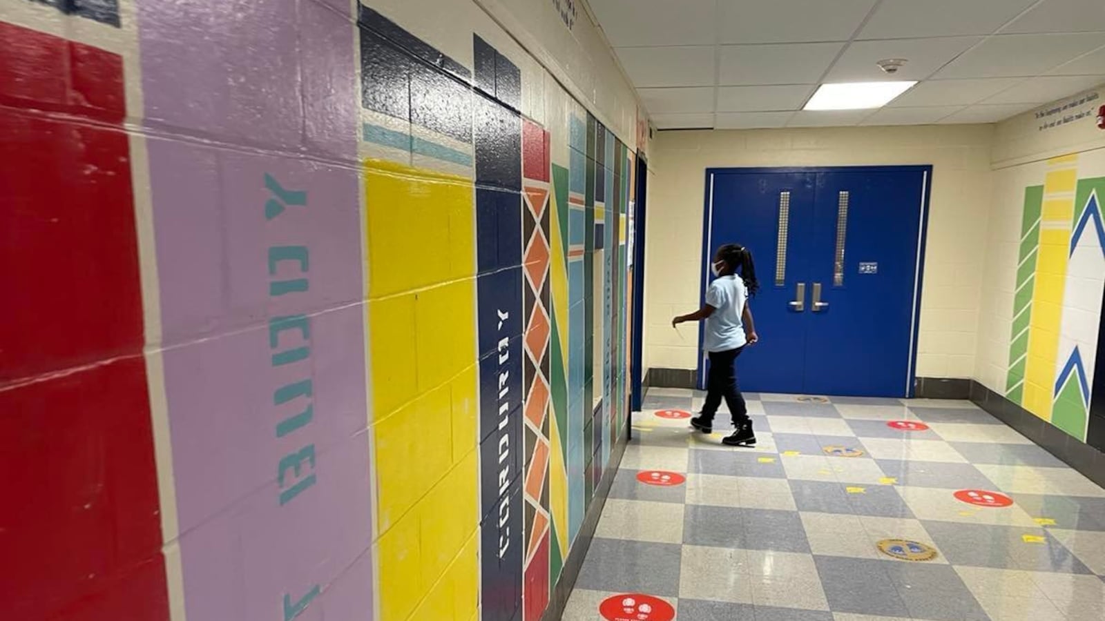A girl in a light shirt and dark pants wearing a face mask walks towards a doorway in a colorful school hallway. 
