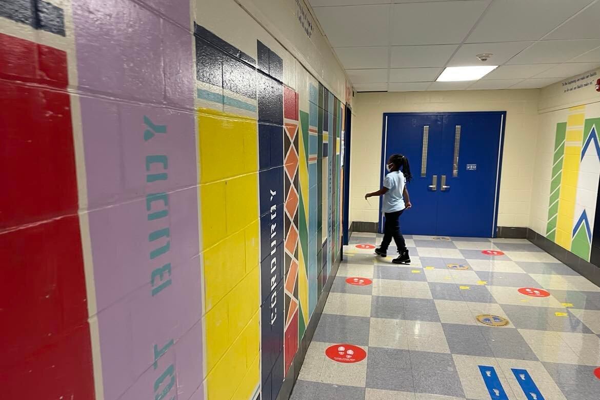 A girl in a light shirt and dark pants wearing a face mask walks towards a doorway in a colorful school hallway.