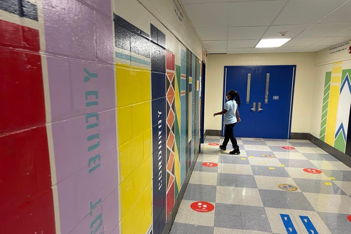 A girl in a light shirt and dark pants wearing a face mask walks towards a doorway in a colorful school hallway. 